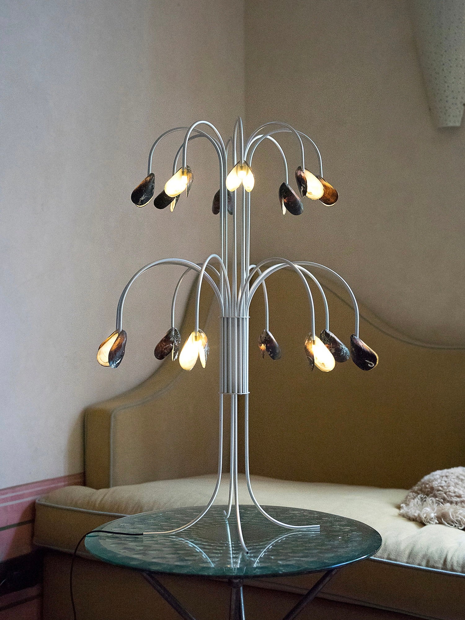 The Shell Light from Touch With Eyes, featuring multiple curved branches each ending in a mother-of-pearl bulb with a shell-like design, is elegantly situated on a round glass table. This handmade lamp casts a warm glow, complemented by the view of a couch in the background against a light-colored wall.