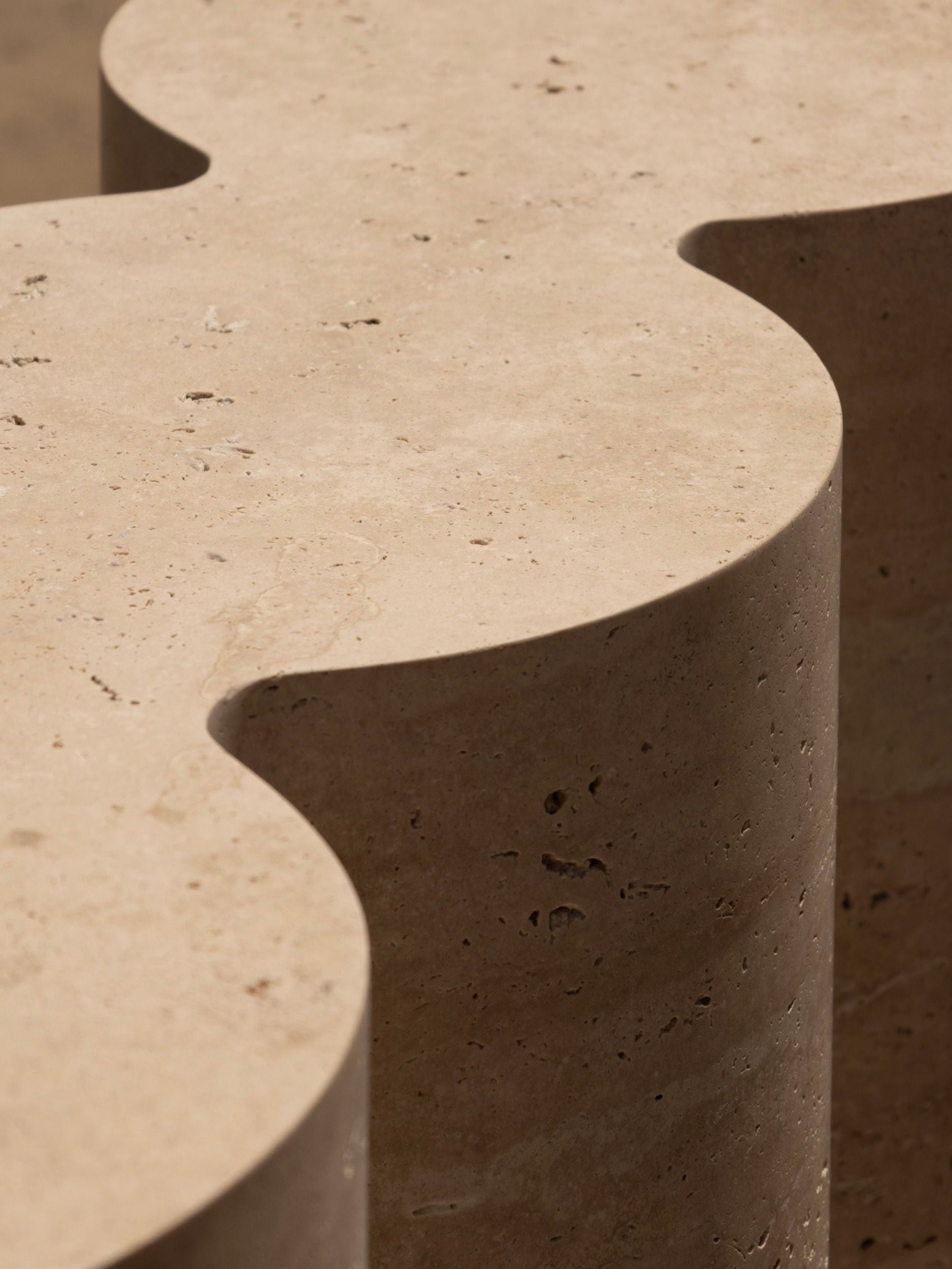 Close-up of the M_008 Travertine Console by Monolith, a beige stone sculpture with a wavy silhouette. Its smooth, textured surface, dotted with small holes, enhances its natural allure. Lighting highlights its curves and edges, casting soft shadows that emphasize this impressive creation.