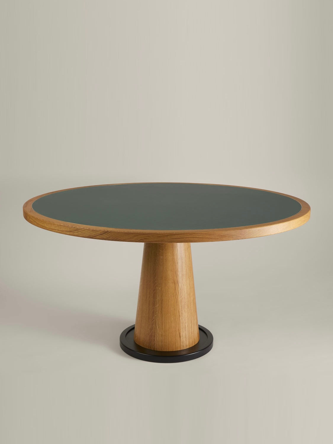 The Jack Table by HAUVETTE & MADANI is a contemporary round wooden table with a sleek dark gray tabletop supported by a tapered cylindrical pedestal. It elegantly sits on a circular black base, reminiscent of tables used in green-carpeted games, and is beautifully set against a backdrop of simple light hues.