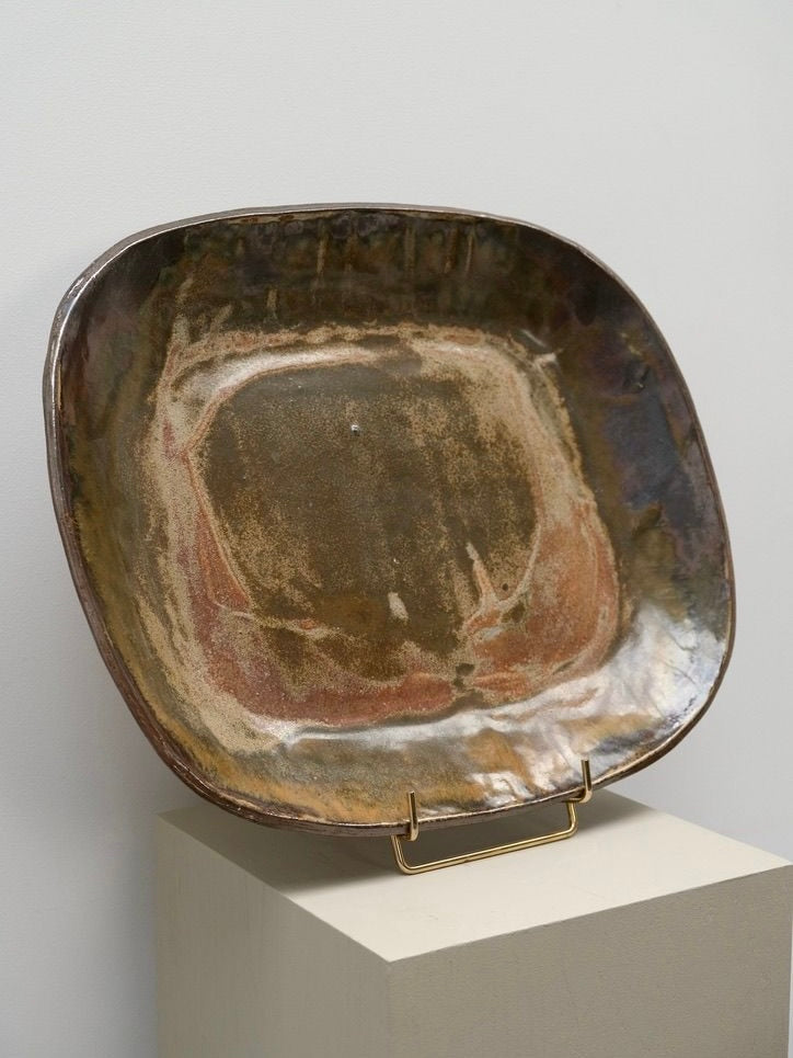 A handcrafted Large Decorative Stoneware Dish by LA NOUVELLE GALERIE with an irregular square shape and a worn, earthy glaze in shades of brown, green, and hints of red. Reminiscent of Pierre Culot's work, this glazed stoneware piece sits on a minimalist display stand atop a rectangular pedestal against a plain white wall.