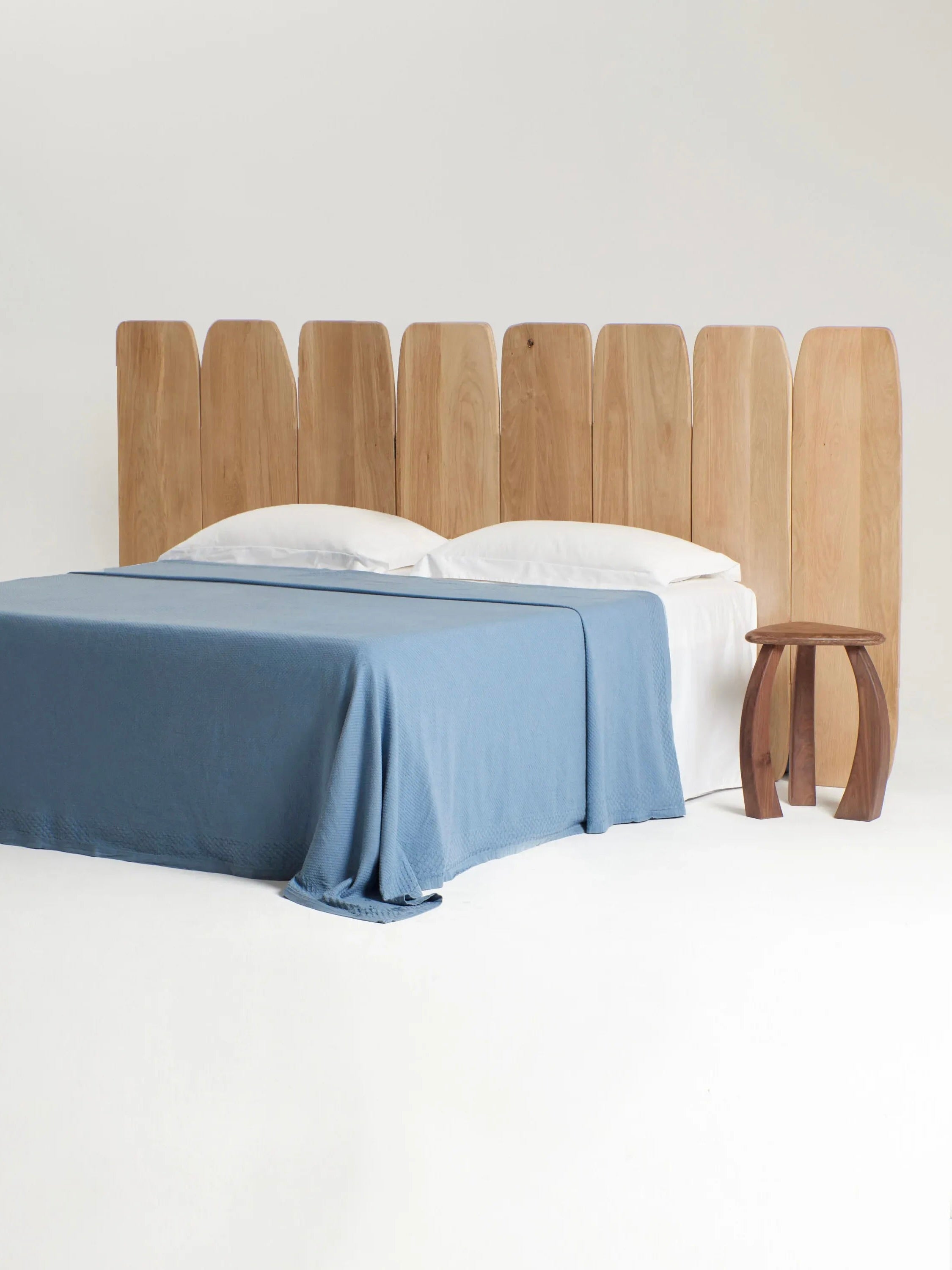 A minimalist bedroom features a wooden bed frame with the Madeira Oak Headboard Medium by Project 213A, made of vertical oak wood planks. The bed is neatly made with a light blue blanket and white pillows. Beside the bed, there is a small, rustic wooden nightstand. The background is white.