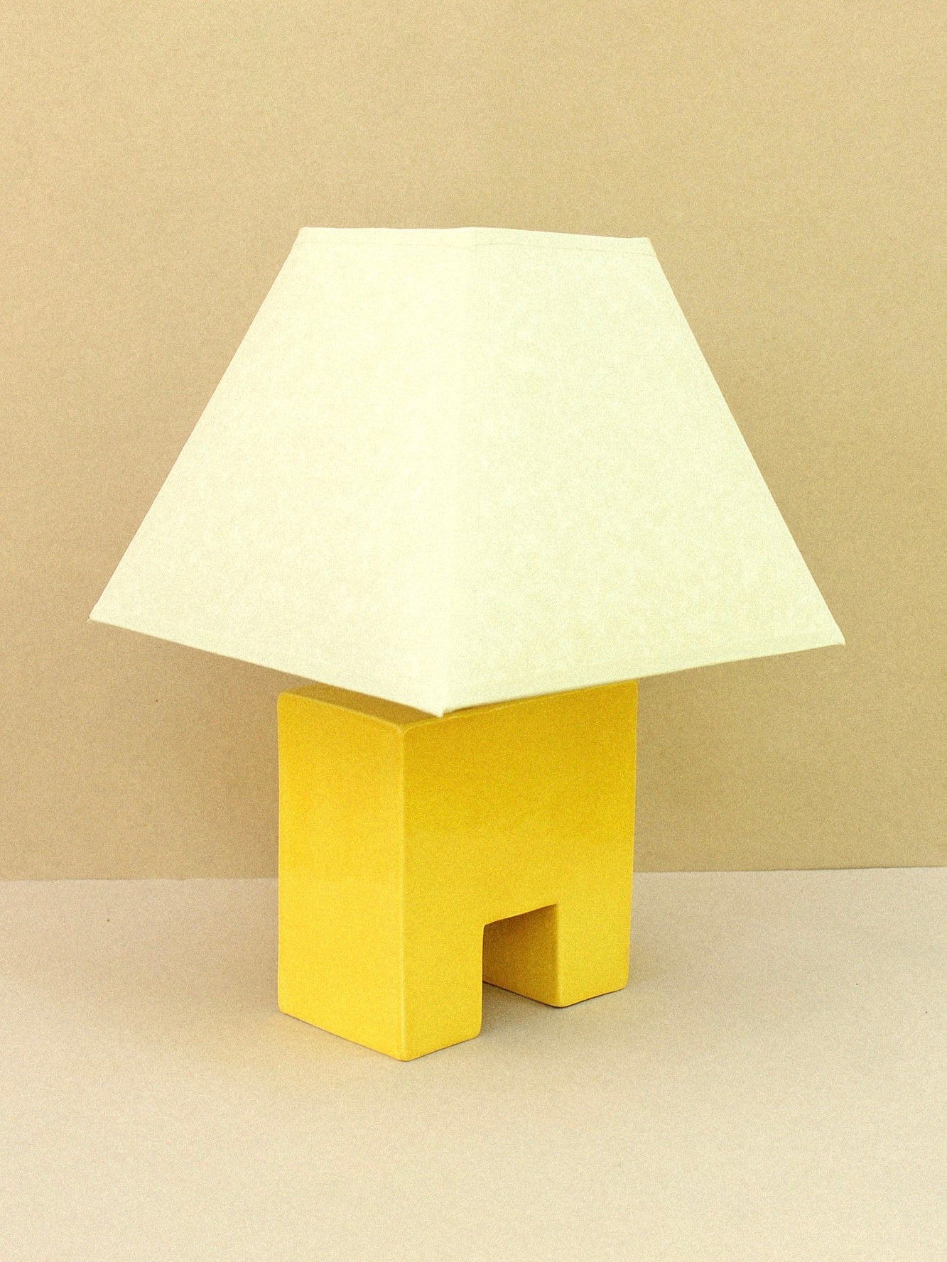 The Cristo Lamp (Sol) by Casa Veronica features a small yellow, rectangular ceramic base and a wide, off-white custom shade, shown against a beige background.