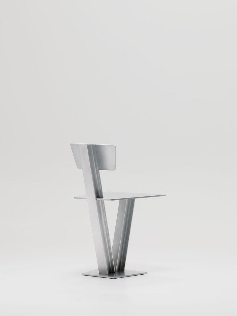 The Beam Chair by Marquel Williams is a modern, minimalist piece featuring a unique geometric design made from waxed aluminum. It has a flat seat, angular backrest, and angled supports against a plain white background.