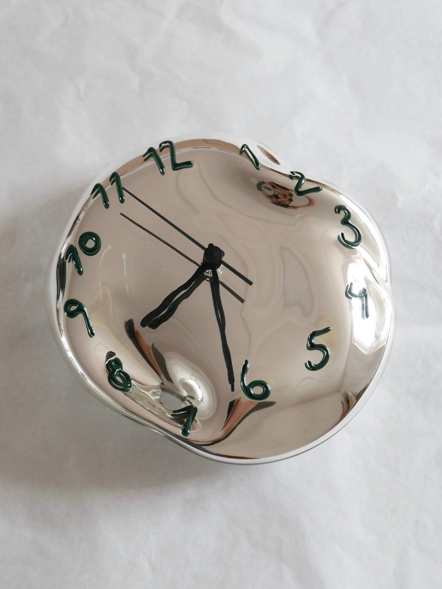 The Silverclock - Green and Black by Silje Lindrup is a handcrafted, distorted reflective clock featuring green numerals and black hands against a crumpled white background. Made in Denmark, this uniquely warped timepiece resembles the surreal artwork of Salvador Dalí while its reliable quartz mechanism ensures accurate timekeeping.