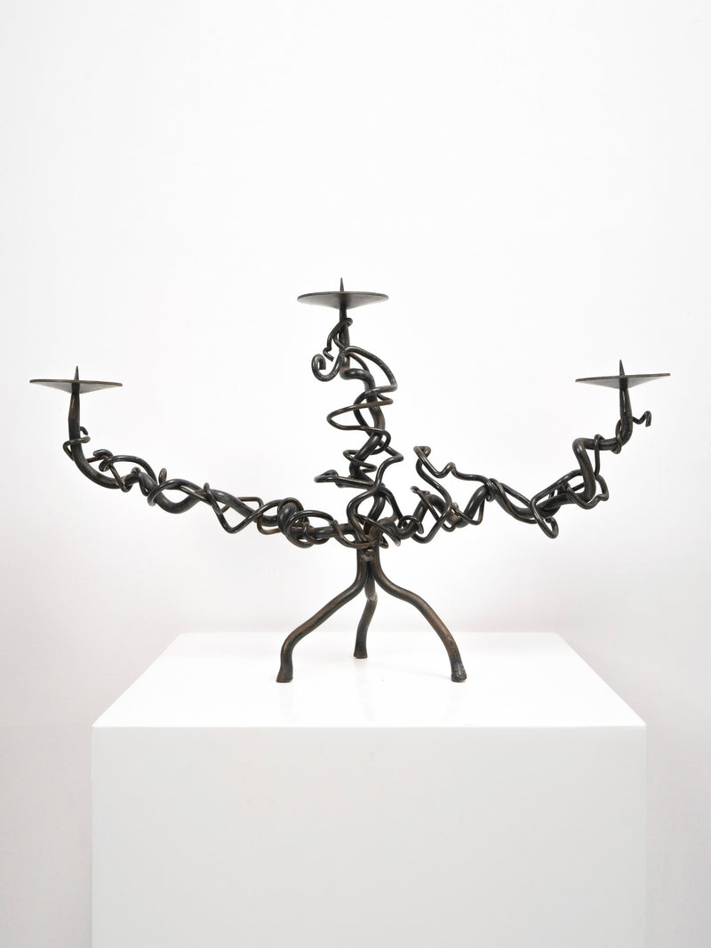The Tom Bogle Brutalist Iron Candelabra features hand-forged, vine-like branches, three candle plates, and tripod legs. Displayed on a white pedestal against a plain background.