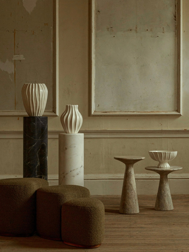 A minimalist interior scene features a ceramic collection with three sculptural vases, including the Large Cloak Vase by Ransom & Dunn, elegantly displayed on tall pedestals. A geometric chair and a compact stone table enhance the textured walls and wooden floor, cultivating an earthy and artistic ambiance. Specialty items crafted by hand in Portugal.