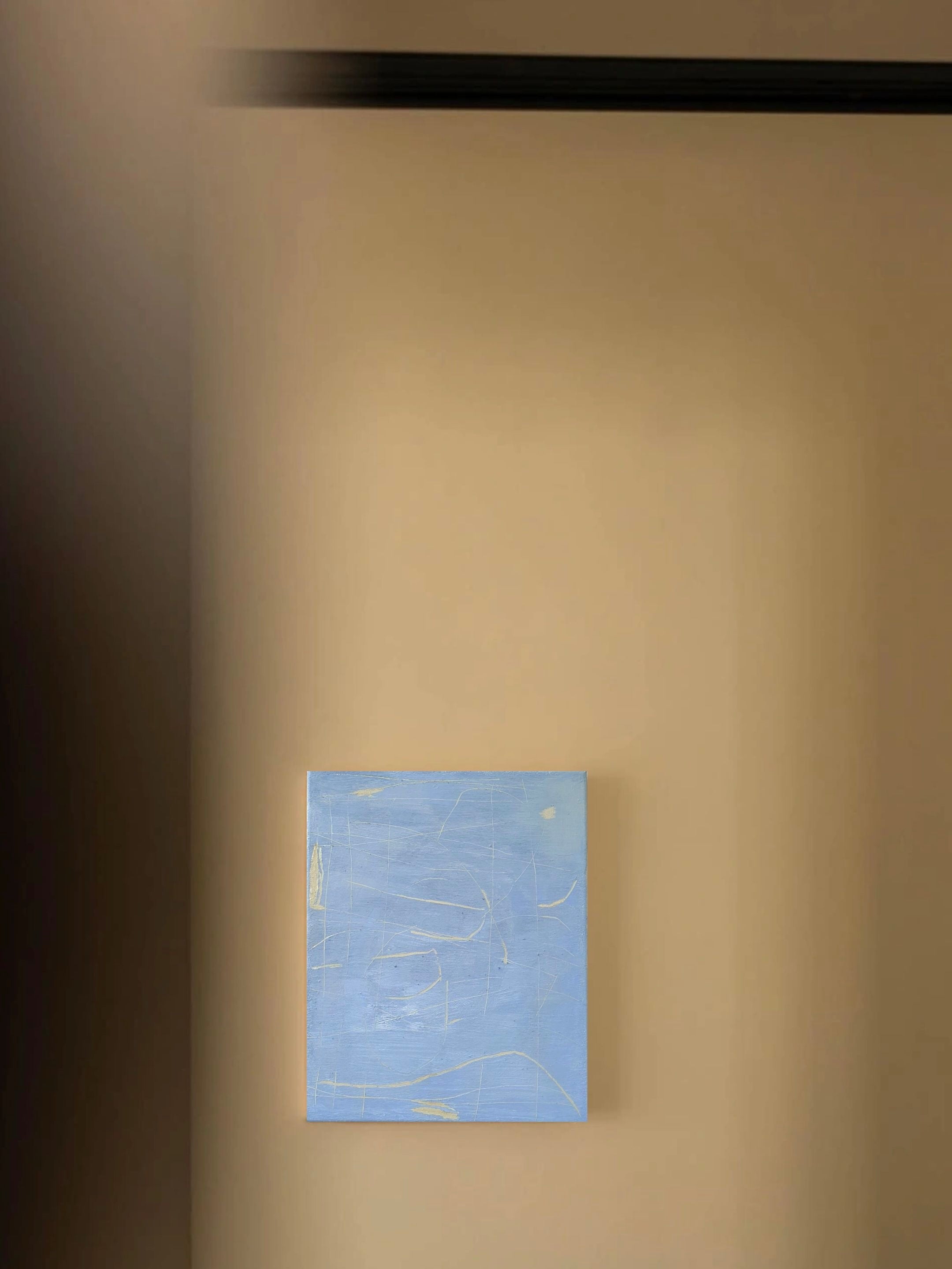 The Unsaid" painting by Yeliza Gevorgyan, featuring faint white and yellow lines on a textured blue canvas, casts a soft shadow on a beige wall. Its minimal marks evoke echoes of ancient worlds, elegantly highlighted by the shadow from an upper black horizontal line.