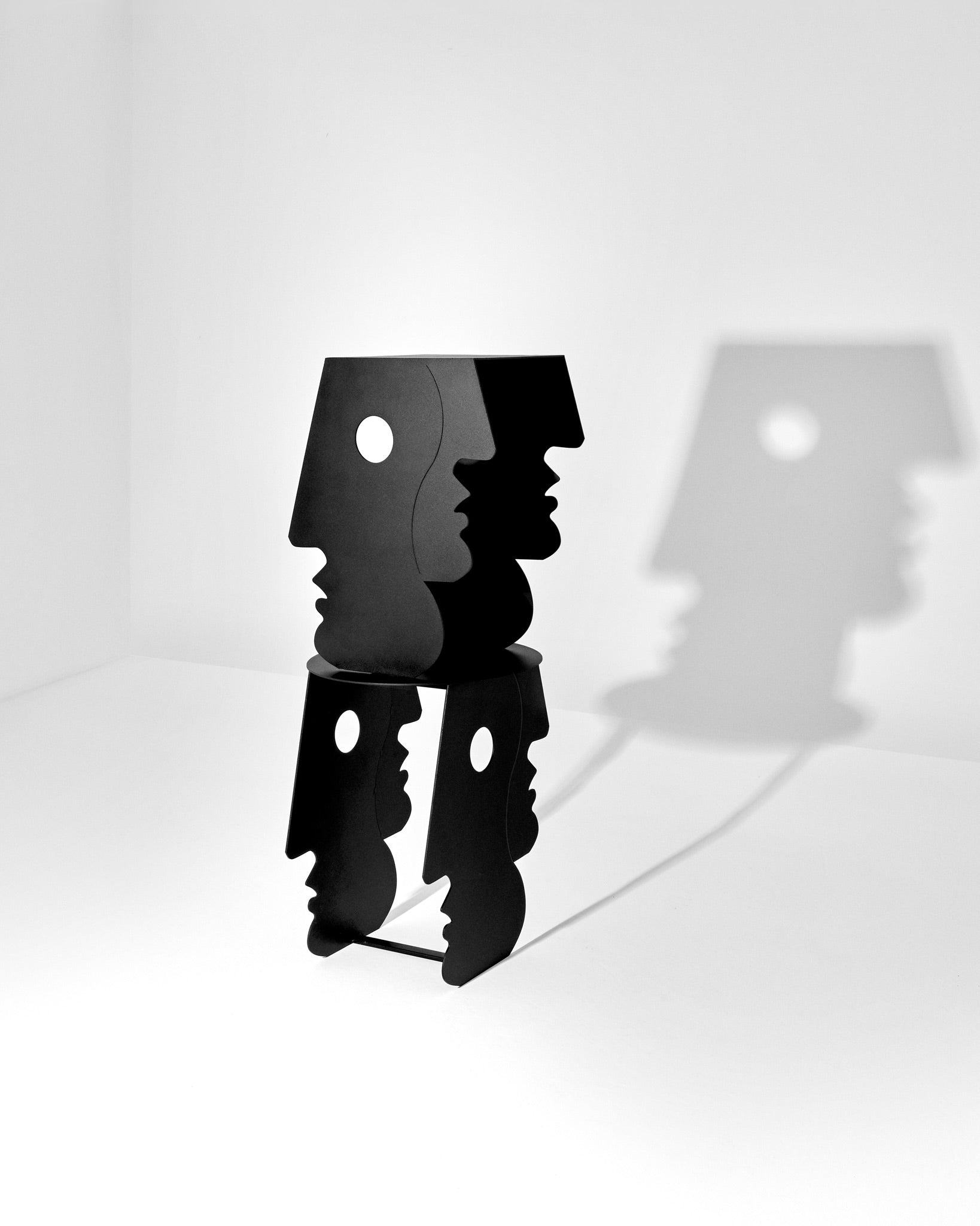The "Lucia" Side Table in Black by Animate Objects, inspired by Bauhaus, stands in a white room, its profile-shaped legs casting shadows on the wall that echo the abstract human forms.