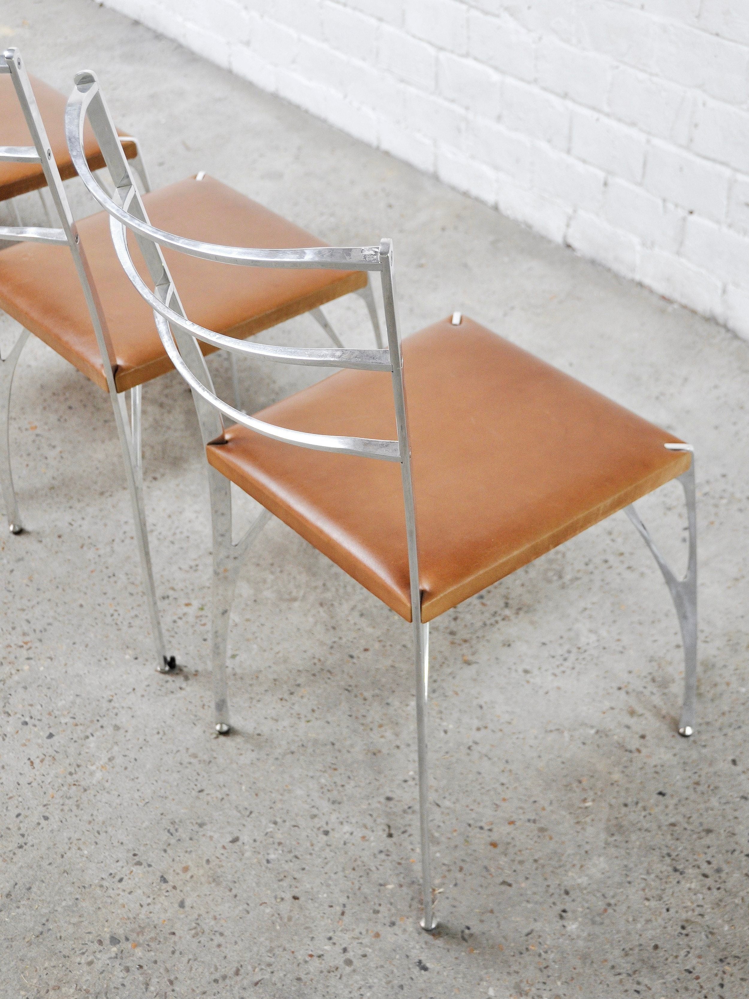 A set of four 'Organic' dining chairs by Erwan Boulloud, from Introverso, with sculptural aluminum frames and brown leather seats rests on a light concrete floor against a white brick wall, showcasing a minimalist and contemporary style.