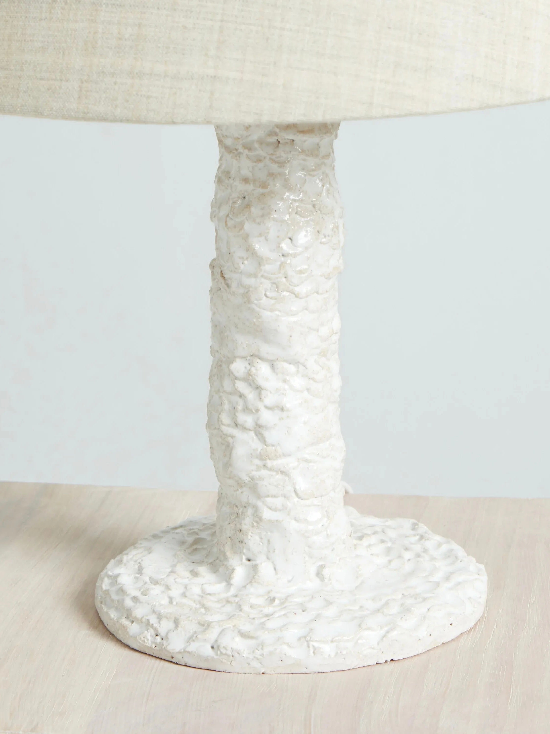 A close-up of the Casa Bedside Light by Project 213A showcases its textured, handmade white ceramic base with a round foundation and cylindrical stem. The fabric lampshade edge is visible, as this elegant piece rests on a light-colored wooden surface.