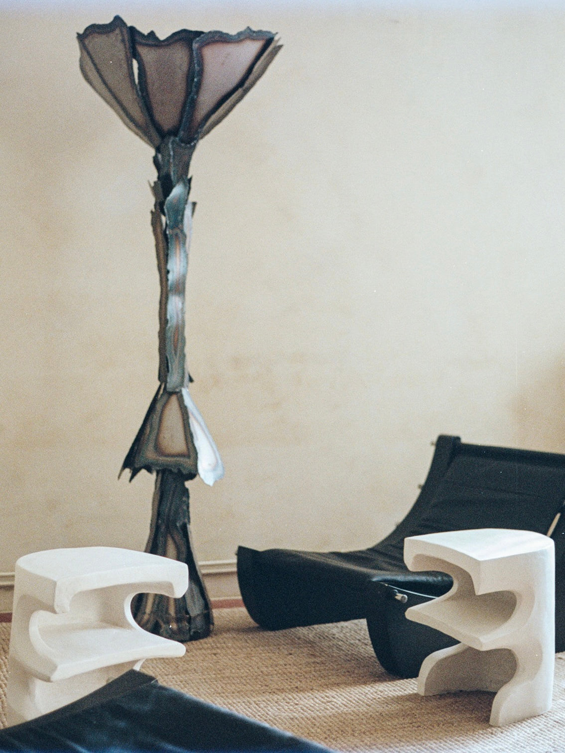 In a contemporary room, a tall metal sculpture stands beside a black lounge chair, complemented by two Baisers Volés Carved Stoneware Coffee Tables from Lea Bigot. These tables rest on a woven rug against a muted wall.