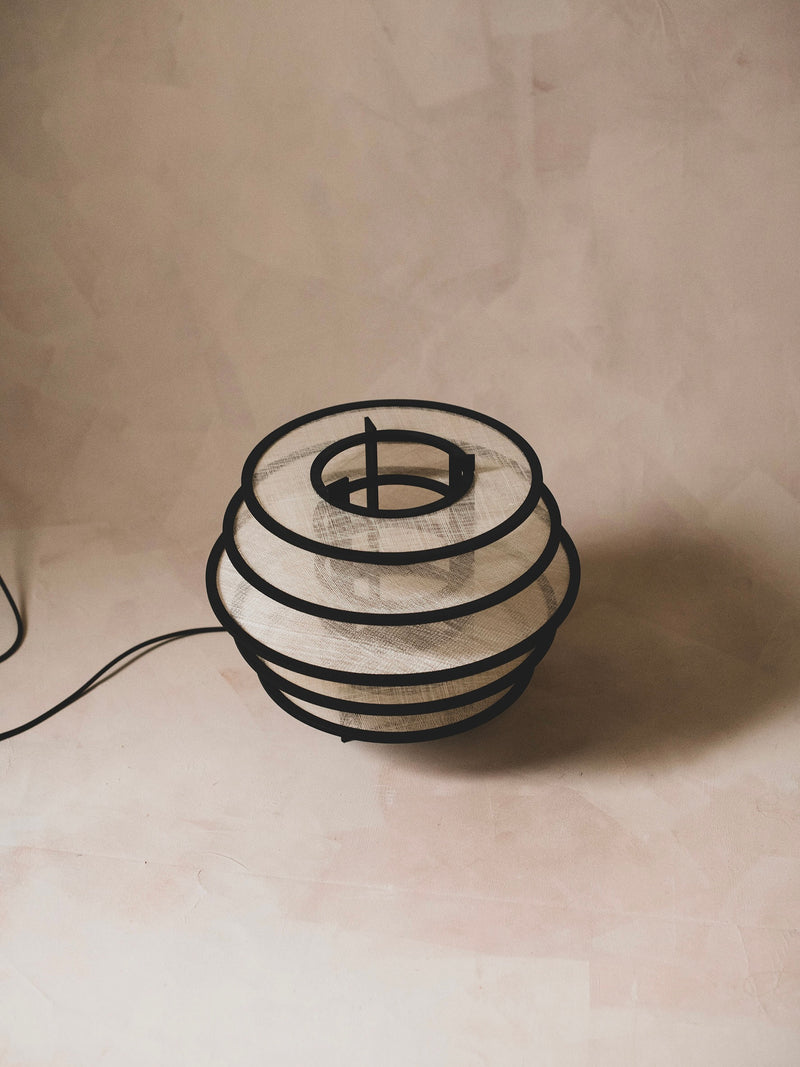 The Lamp I by ELIURPI showcases a sculptural, minimalist design with concentric black metal rings and a textured white shade, providing ambient light against a soft, neutral background.