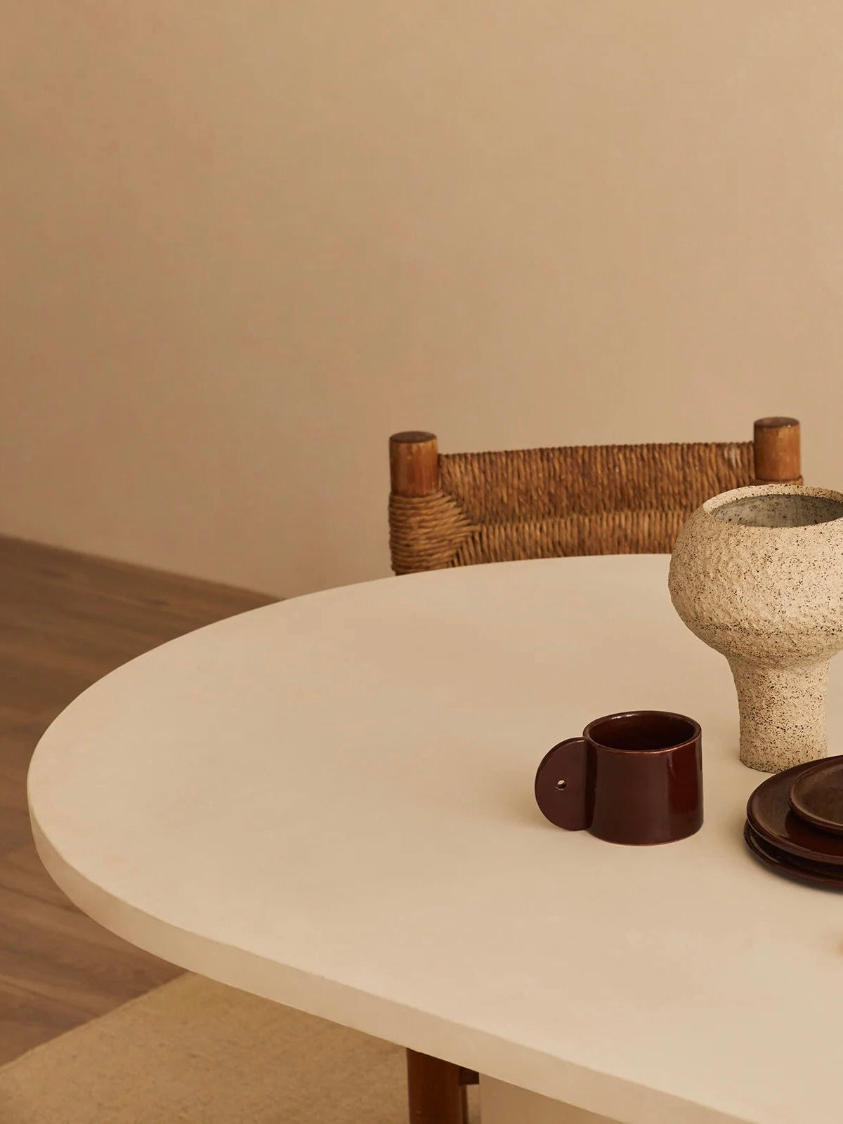 A minimalist scene showcases the MarlotBaus Reye Dining Table, handcrafted in round white microcement. It features a brown ceramic mug, beige textured vase, and dark coasters. A woven chair against a neutral wall complements the contemporary dining furniture style.