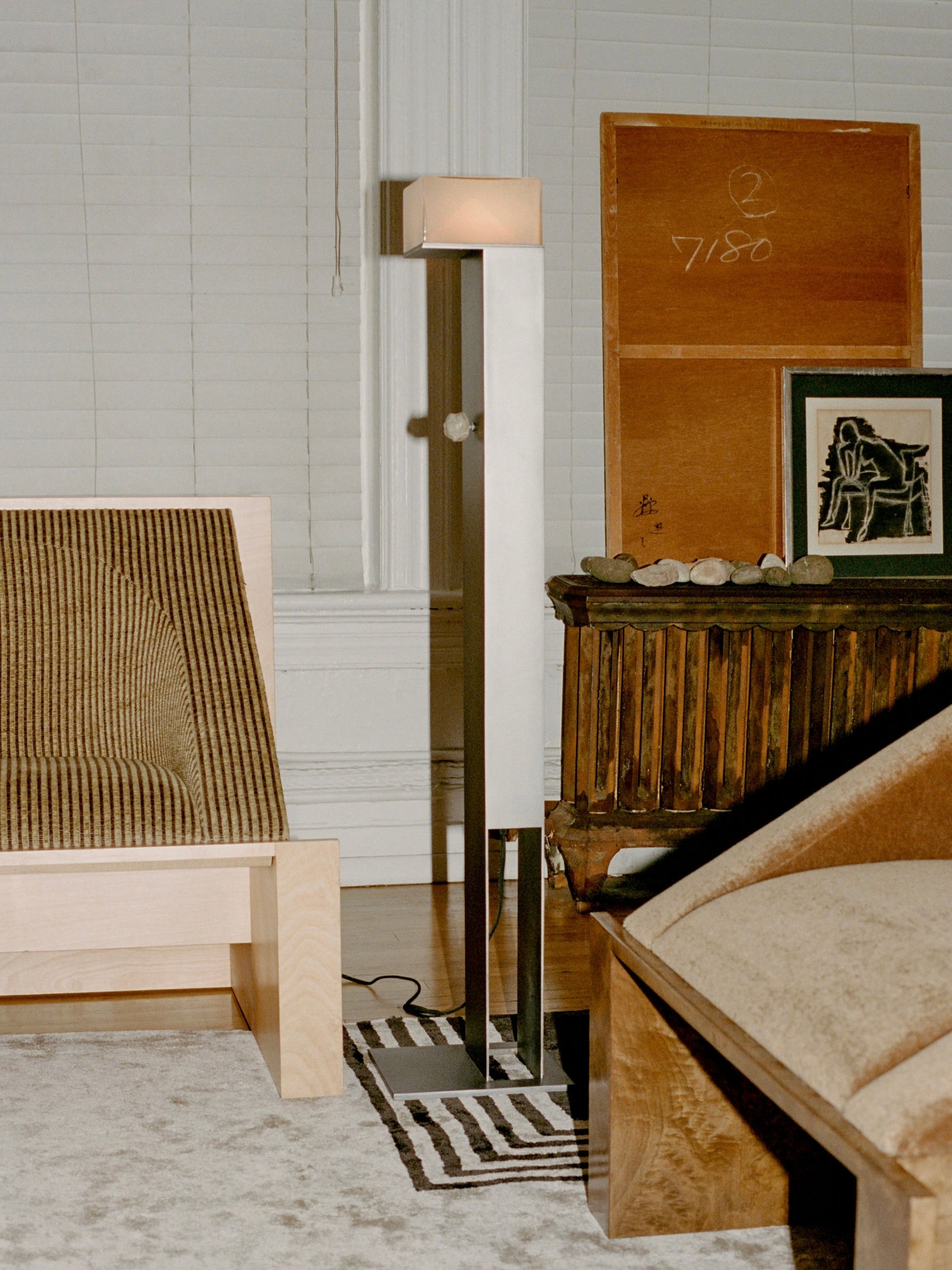 A modern living room embodies minimalist values with a sleek floor lamp beside a striped rug. A 'Perceptions' Sofa Bench by Known Work adds sophistication, along with two wooden chairs with brown cushions, paintings, and a framed black-and-white portrait. Closed blinds complete this serene space.