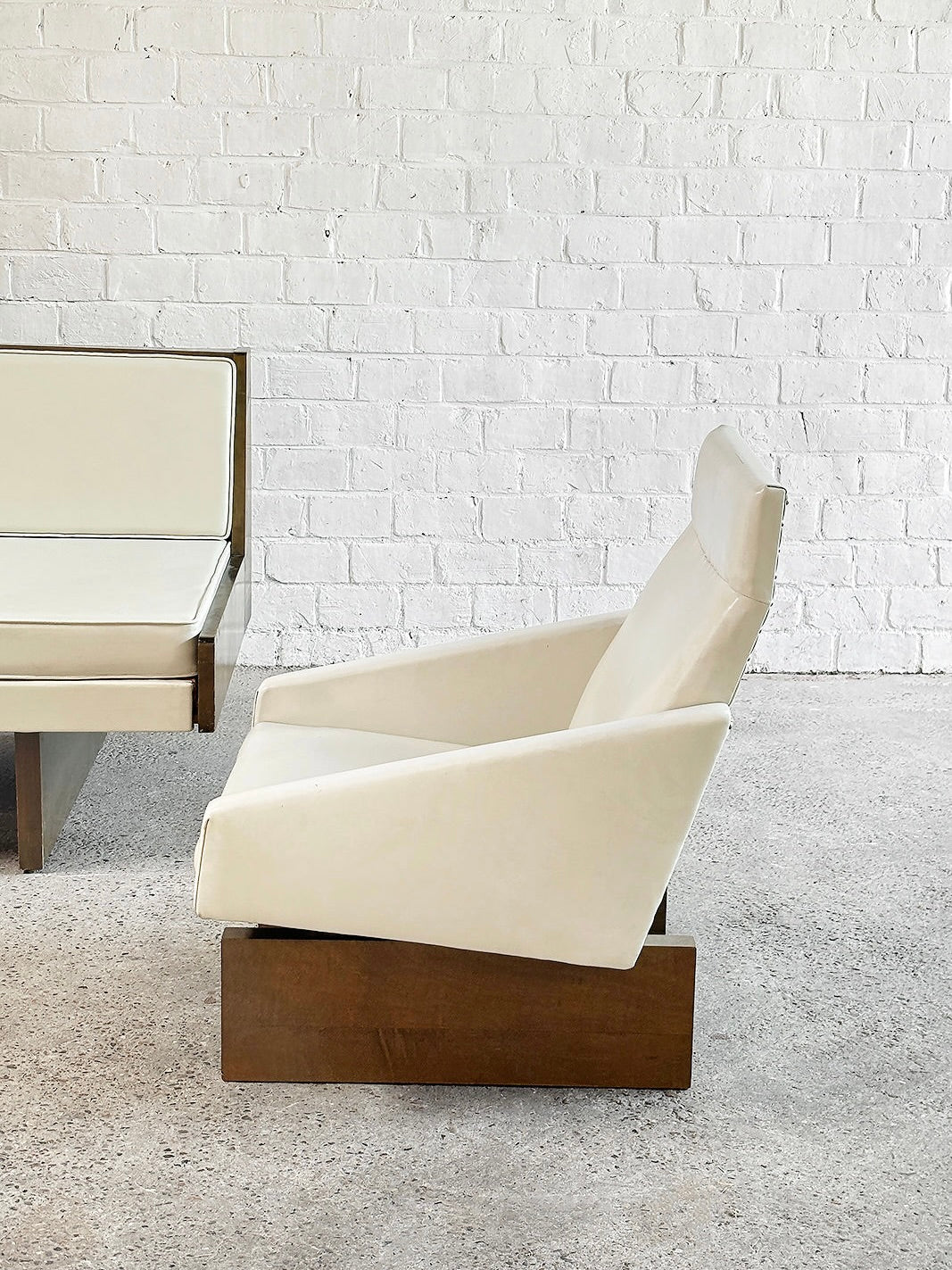 A white modern chair with a wooden base and eco-leather upholstery stands against a white brick wall, next to part of an Introverso's 1950s French Modernist Sofa Set in White Leather. The minimalist décor complements the light floor, merging elegance and simplicity gracefully.