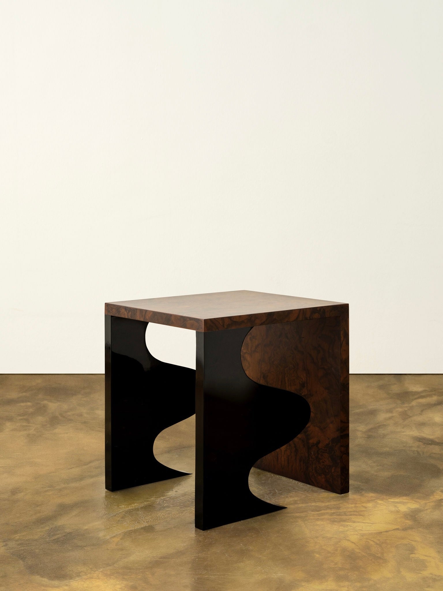 Against a plain white wall, the Harlequin Side Table from Animate Objects rests on a polished concrete floor. This modern, limited edition piece showcases a handcrafted walnut burl veneer top with gloss lacquered wood and uniquely curved black legs. It features artistic wavy cutouts on each side, epitomizing luxury furniture design.