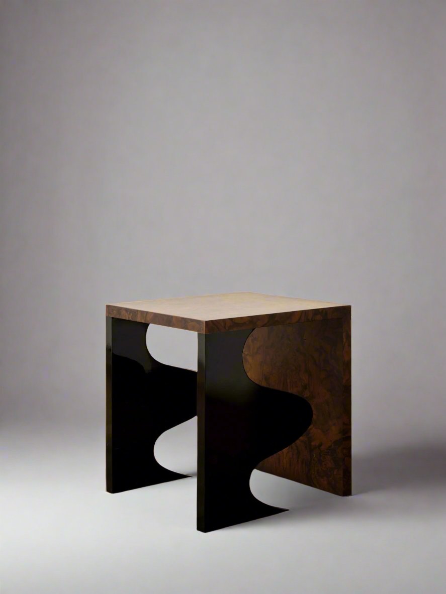 Against a plain white wall, the Harlequin Side Table from Animate Objects rests on a polished concrete floor. This modern, limited edition piece showcases a handcrafted walnut burl veneer top with gloss lacquered wood and uniquely curved black legs. It features artistic wavy cutouts on each side, epitomizing luxury furniture design.