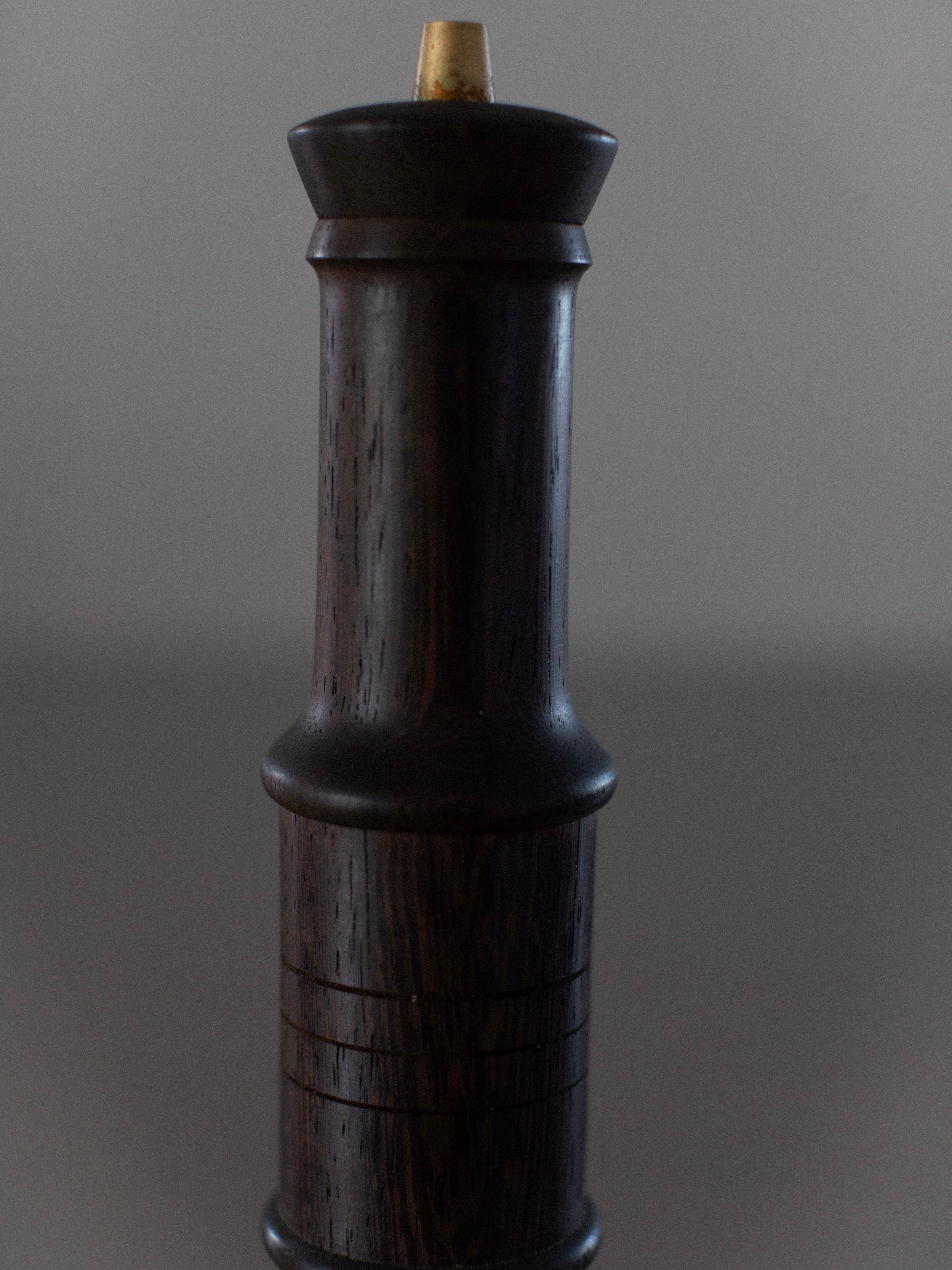 A close-up shot of the Wenge Wood Grinder by Out For Lunch, reminiscent of a vintage Danish pepper grinder. Crafted from wenge wood, it boasts a cylindrical shape with a smooth, polished surface and subtle grain patterns. A brass knob adorns the top, and the background is plain and slightly out of focus.