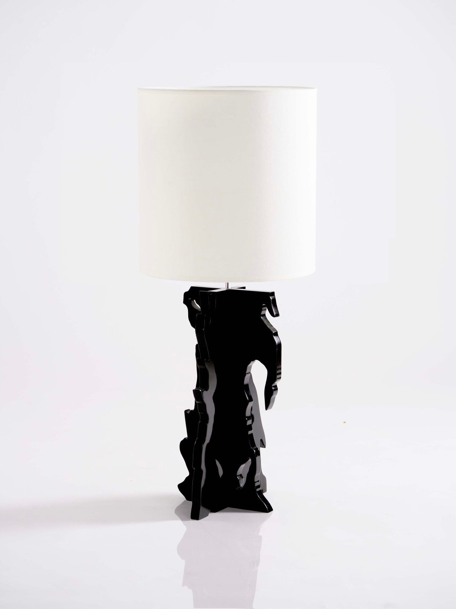 A contemporary lighting piece, "The Bird" Table Lamp by Ochoa Rocabert, features a glossy black base with an abstract design reminiscent of conceptual art. It is complemented by a cylindrical white lampshade set against a plain white background, highlighting the sophisticated allure of lacquered wooden forms.