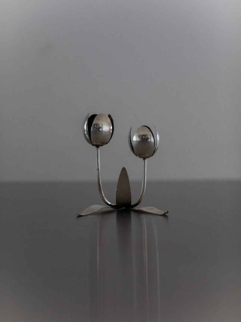 The Steel Tulip Candleholder by Out For Lunch is a captivating piece of modern home decor. This unique metal sculpture, resembling two spoon-like flowers, showcases rounded forms akin to petals or seed pods in a sleek steel design. It features a central stem with a sturdy base set against a soft gray background.