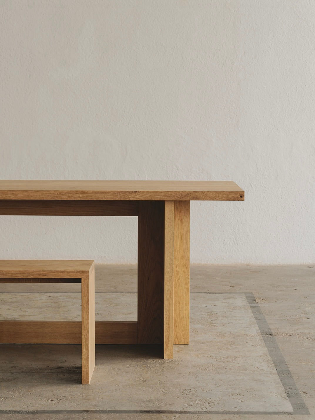 The Paris Dining Table by ASERIES OF OBSTACLES, SL, and its minimalist wooden bench sit gracefully on a textured concrete floor against a light-colored wall. Perfect for intimate gatherings, the set features clean lines and a natural wood finish.