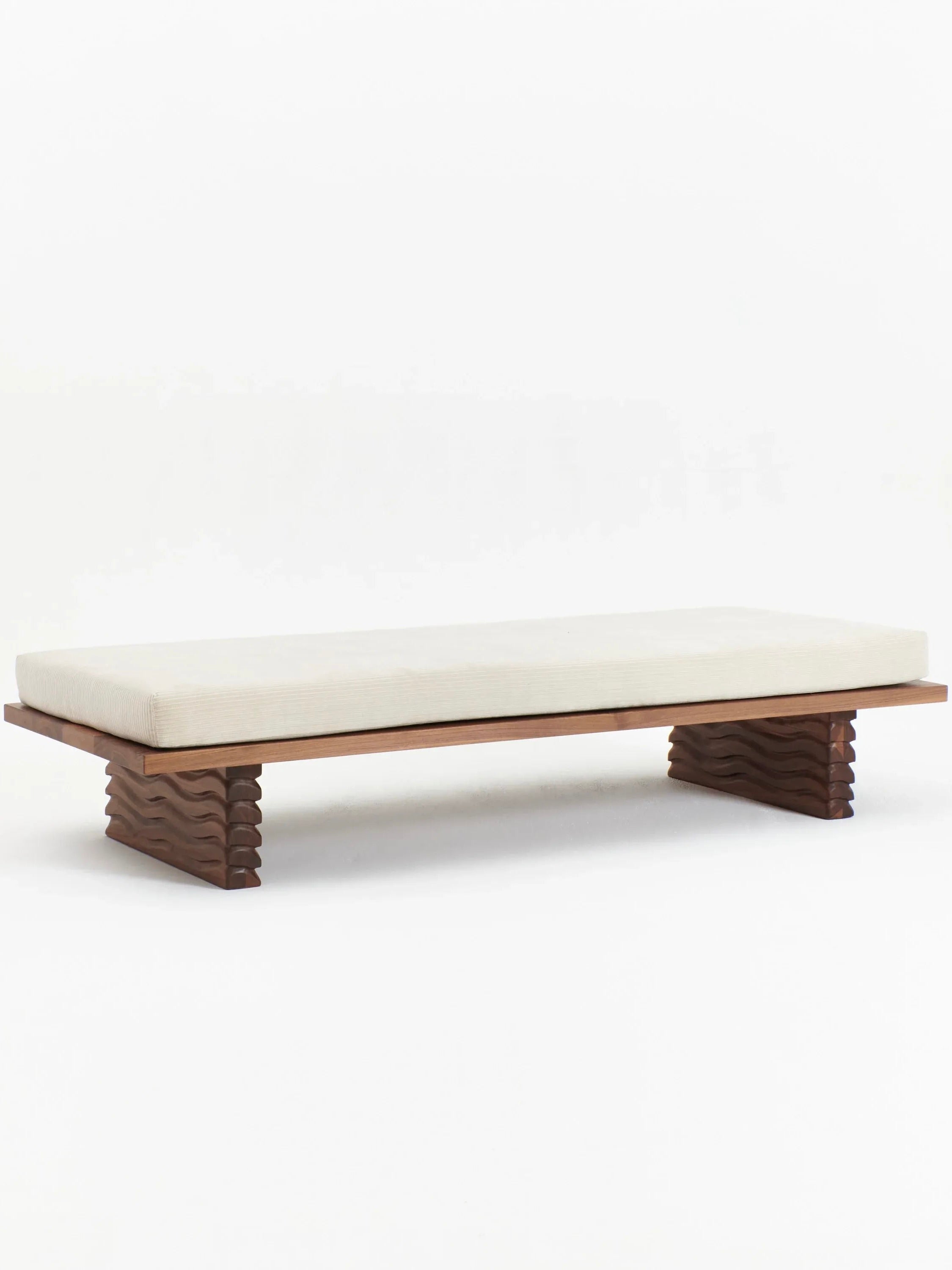 Introducing the Nazaré Daybed by Project 213A: a minimalist bench, handmade in Portugal, featuring a light beige cushion atop a solid walnut base. The intricately carved wave-like patterns on its legs evoke an artistic and contemporary design. The daybed is set against a plain white background.