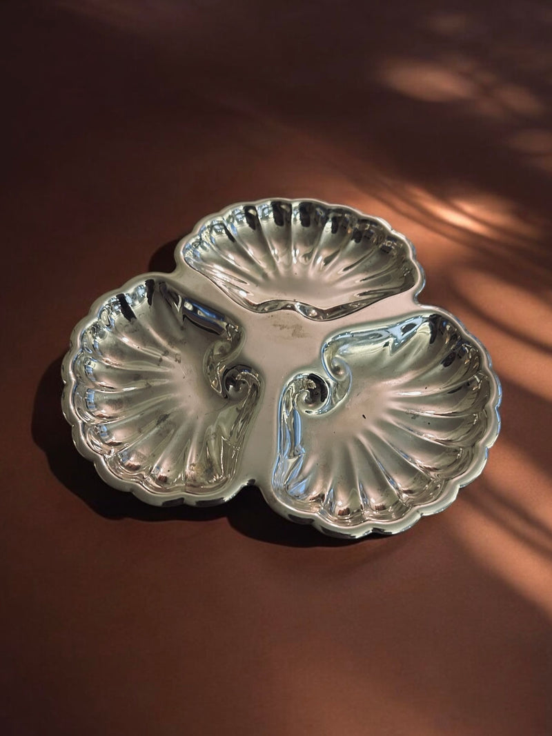 The Flock Studio Triple Shell Server is a silver piece with three scalloped, shell-like compartments set against a soft brown background featuring gentle light patterns.