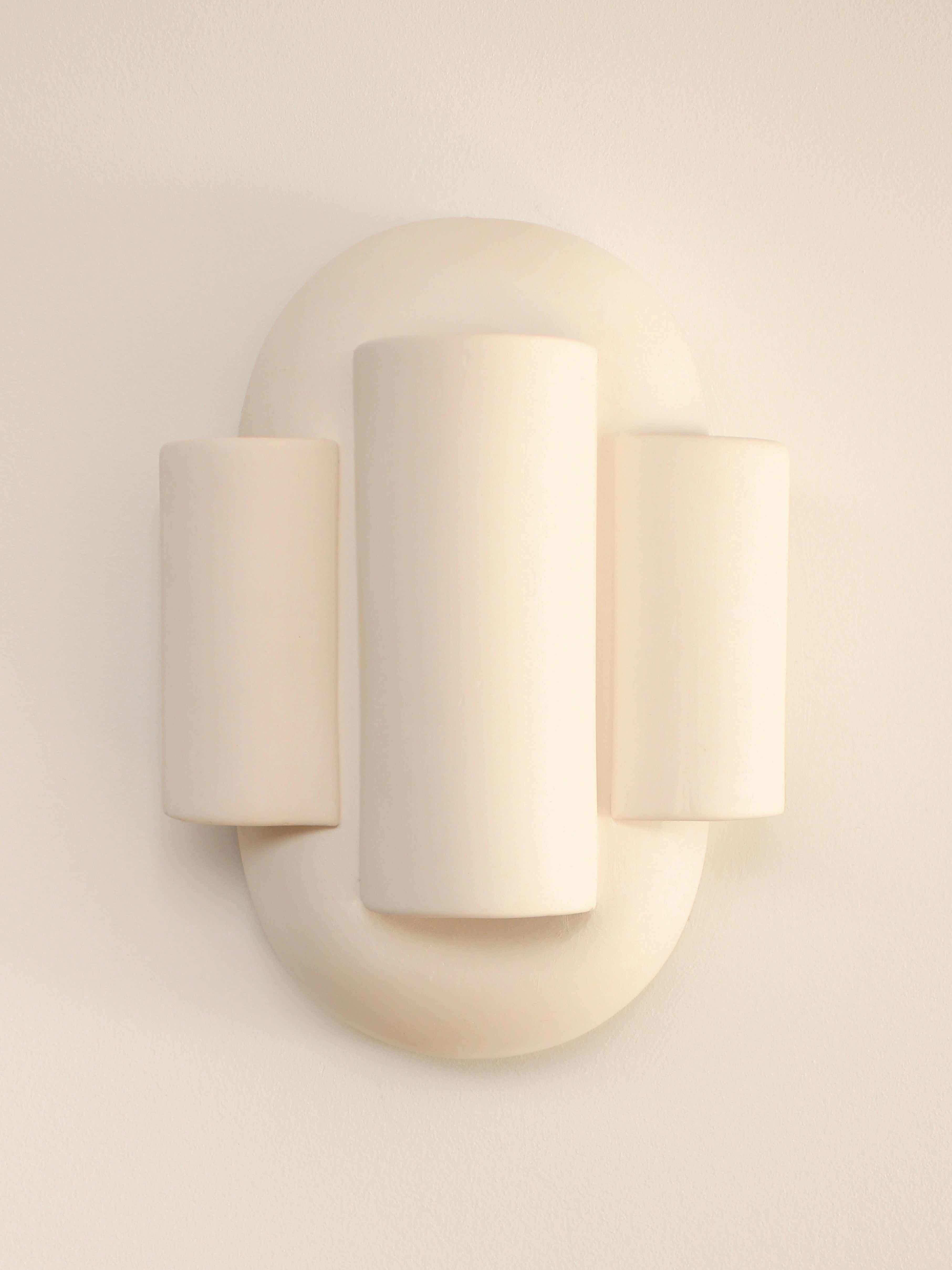 The YEREK Wall Light by Villa Arev is a sculptural wall fixture with a minimalist, modern design. It features three cylindrical forms in soft white, with the central largest cylinder flanked by two smaller, equally sized cylinders on a rounded rectangular base. The muted tones convey an elegant simplicity suitable for any existing socket.