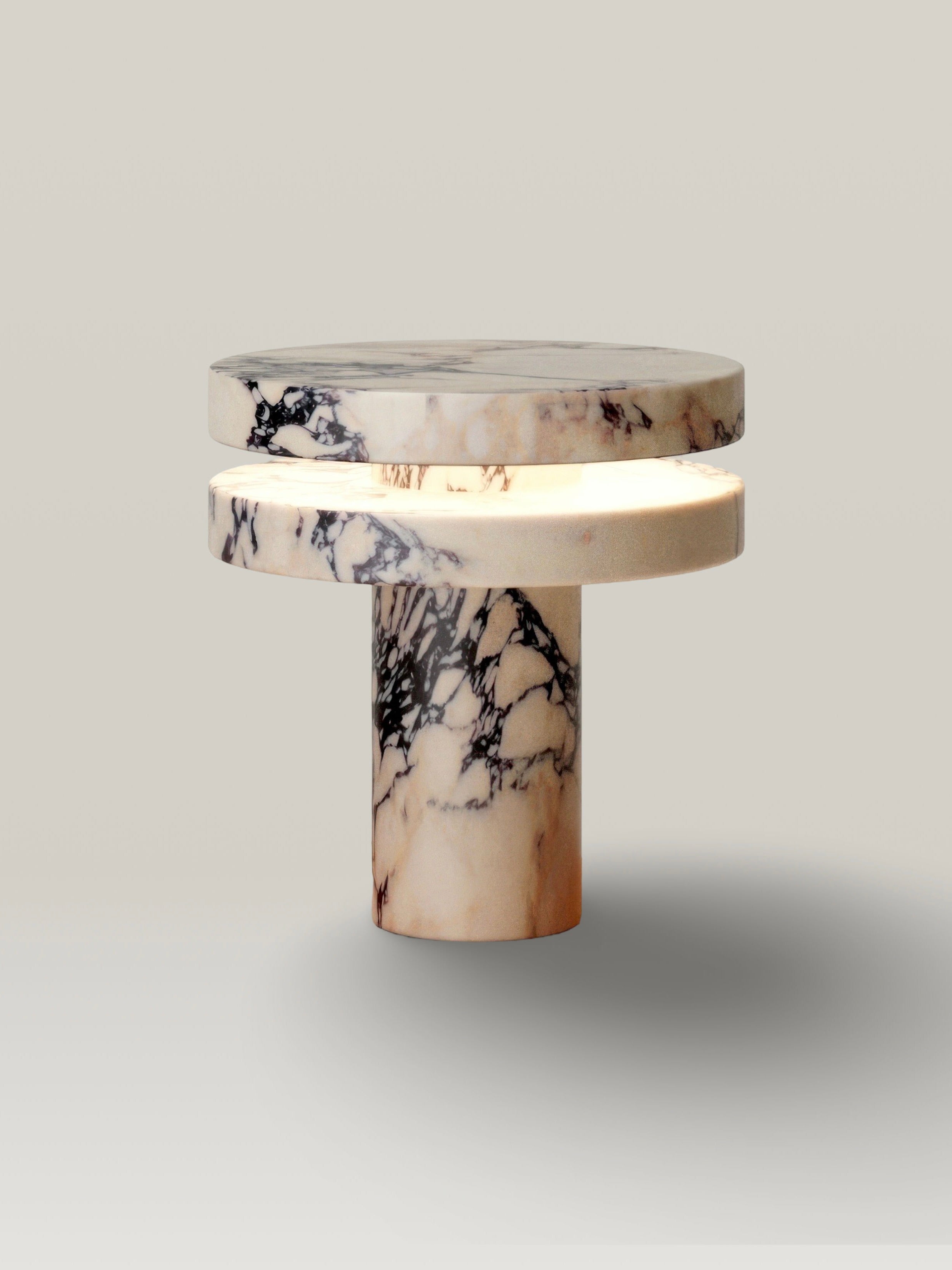 The 'M_014' Marble Table Lamp by Monolith features a minimalist, elegant design with a cylindrical base and two stacked, marble-like circular layers. The top layer illuminates with a dimmable LED, casting a soft glow against any neutral background.