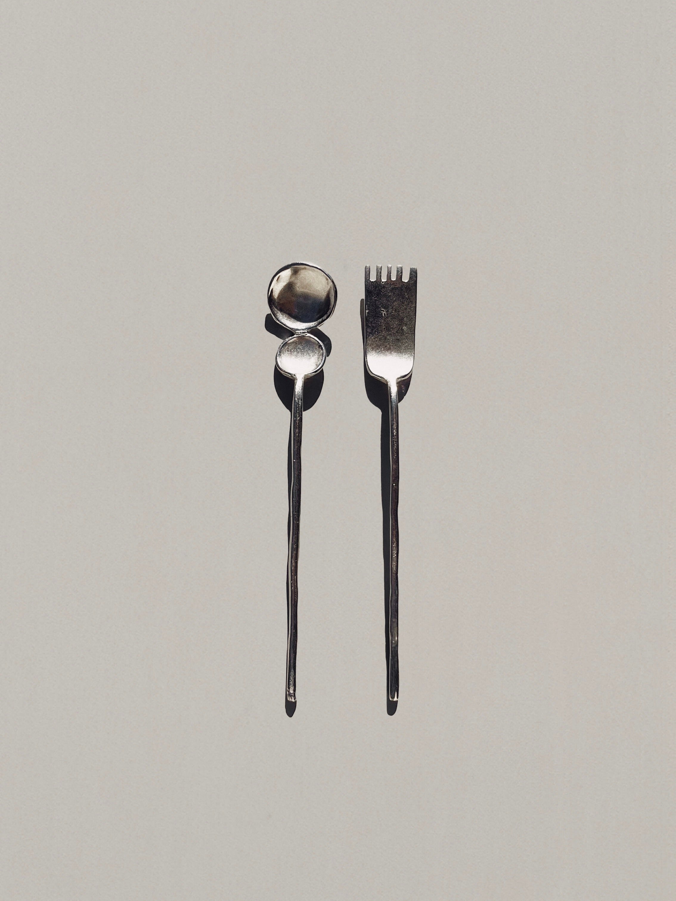 The "Artifact" Tea Cutlery - Duo by Hors-d'Oeuvre consists of a spoon and fork with elongated, twisted handles that exude a mystical aura against a light gray background. The lighting highlights their metallic surfaces, echoing the charm of ancient ornaments.