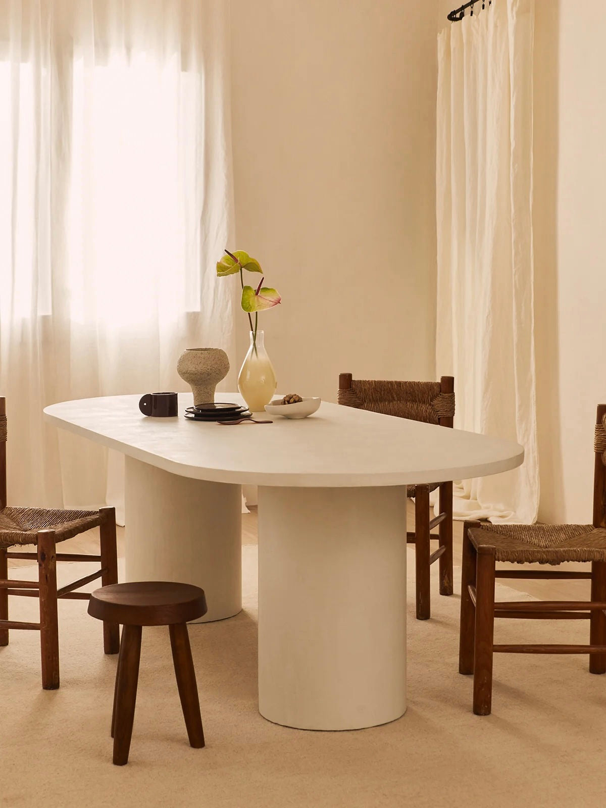 A minimalist dining room highlights the MarlotBaus Reye Dining Table, accompanied by wooden chairs and a stool. The table is adorned with a vase of flowers, a bowl, a mug, and decor. Soft natural light softly diffuses through sheer curtains.