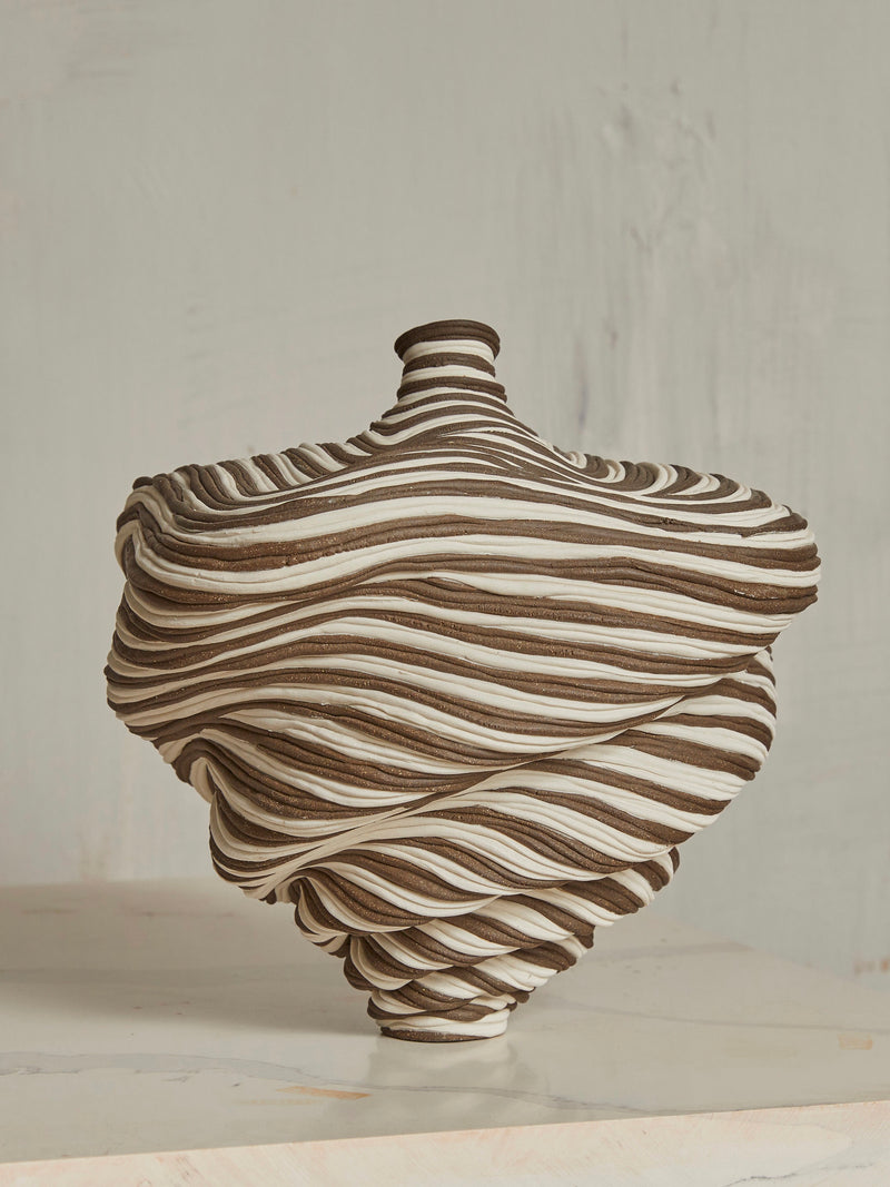The Soucy Shop's Black & White Bottle, a hand-rolled porcelain vase with a spiral pattern of brown and white stripes, features an asymmetric twist, flaring outward before narrowing at the neck. It sits on a marble surface against a light textured wall that complements its artistry.