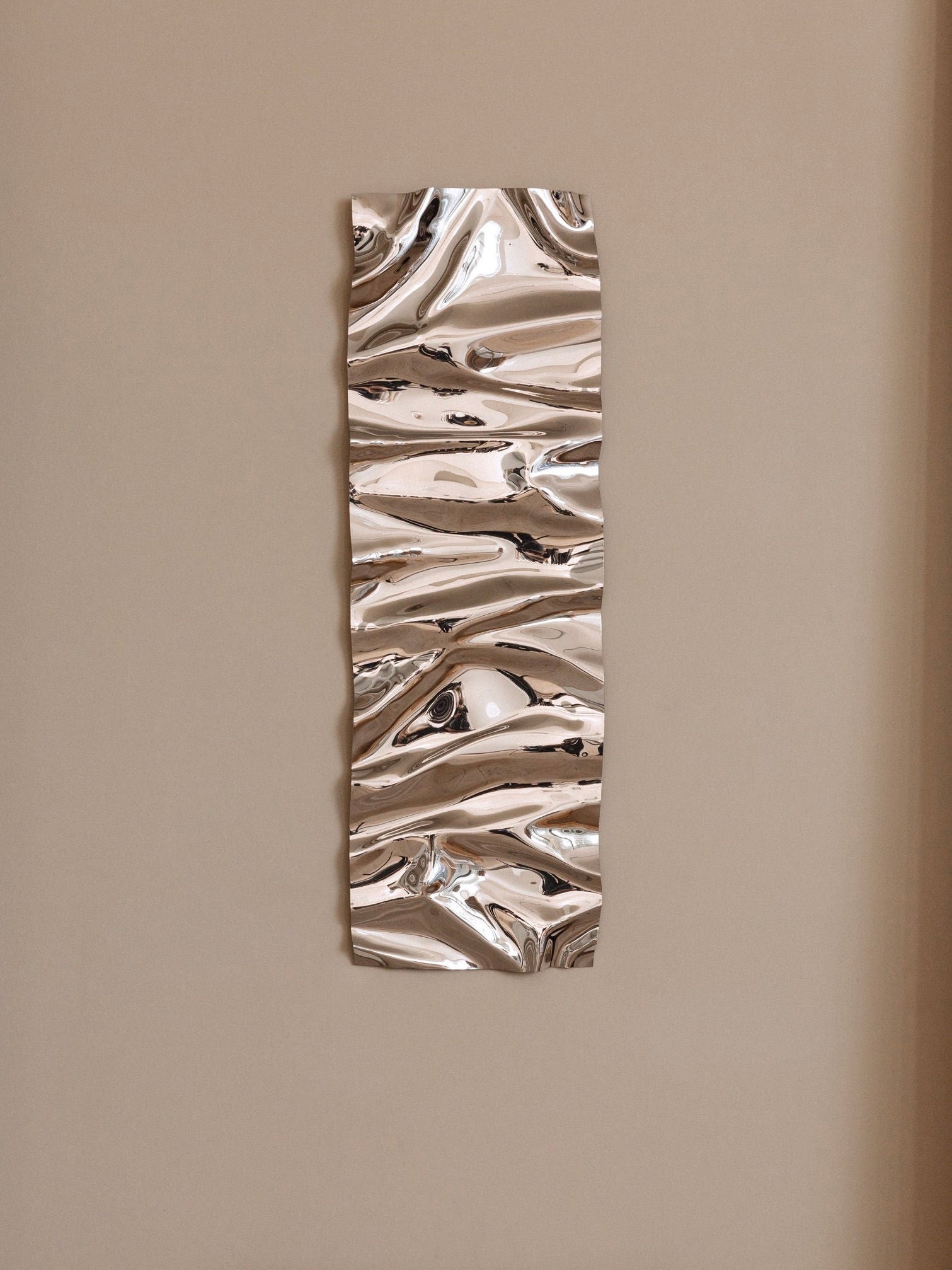 Introducing the *Psychedelic Mirror Long* by *Caia Leifsdotter*, a wall-mounted, rectangular metallic sculpture showcasing an abstract, wavy design. Its reflective surface crafts dynamic light patterns across the textured, rippled features. This handmade sculptural mirror is elegantly displayed against a plain beige background.