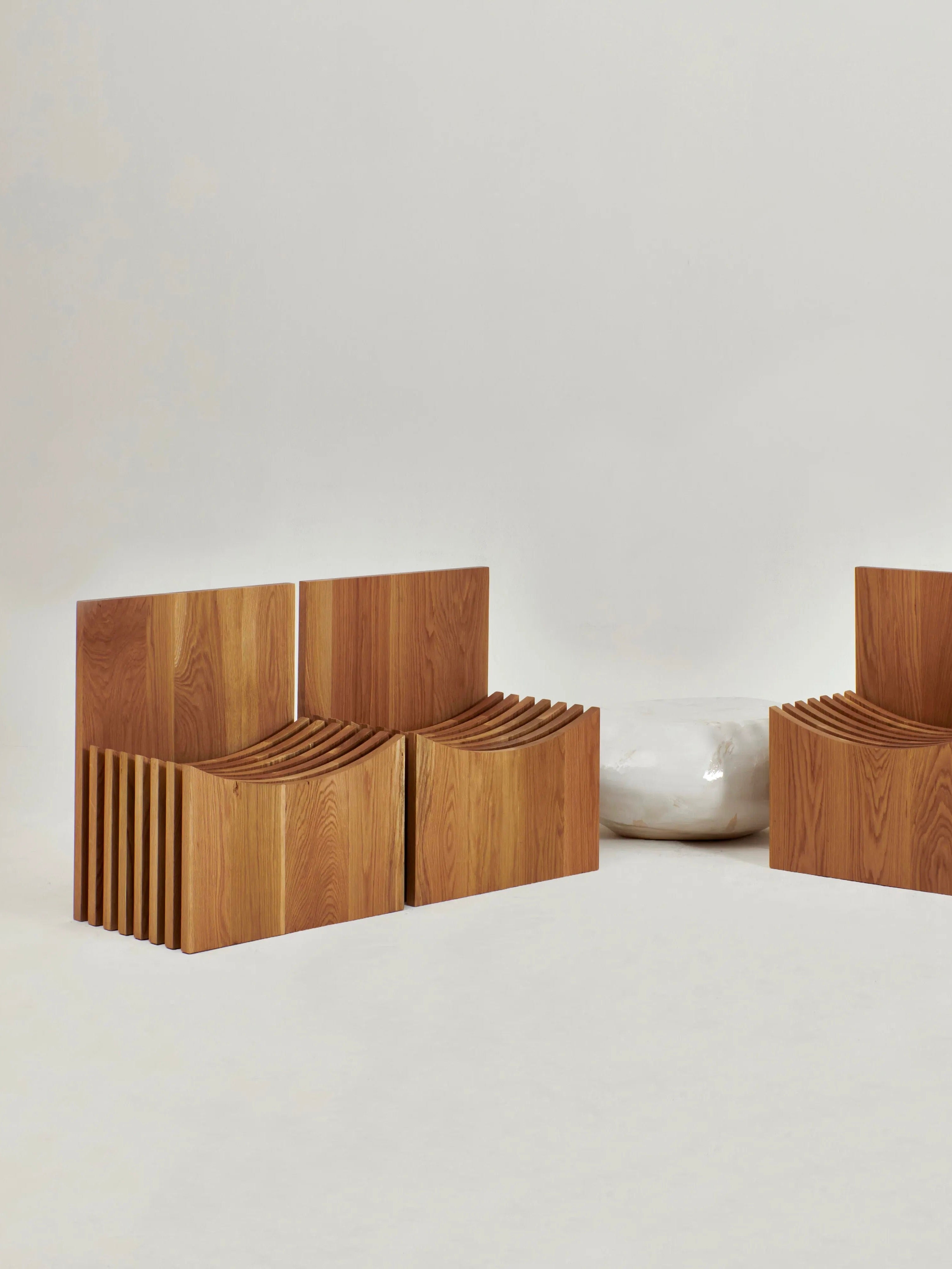 The image showcases two wooden Melides Chairs by Project 213A, each boasting a modern slatted design, positioned facing each other. In between, a minimalist white round cushion or pouf rests on a simple backdrop. The solid oak frames of the chairs underscore their hand-crafted quality and enhance the overall aesthetic.