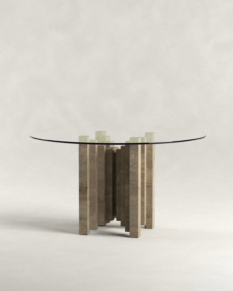 The 'Calakmul' Dining Table S2 by Siete Studio features a modern round glass top supported by a unique travertine base with handcrafted stone pillars arranged circularly. The background is a neutral, light gray, enhancing its elegant design.