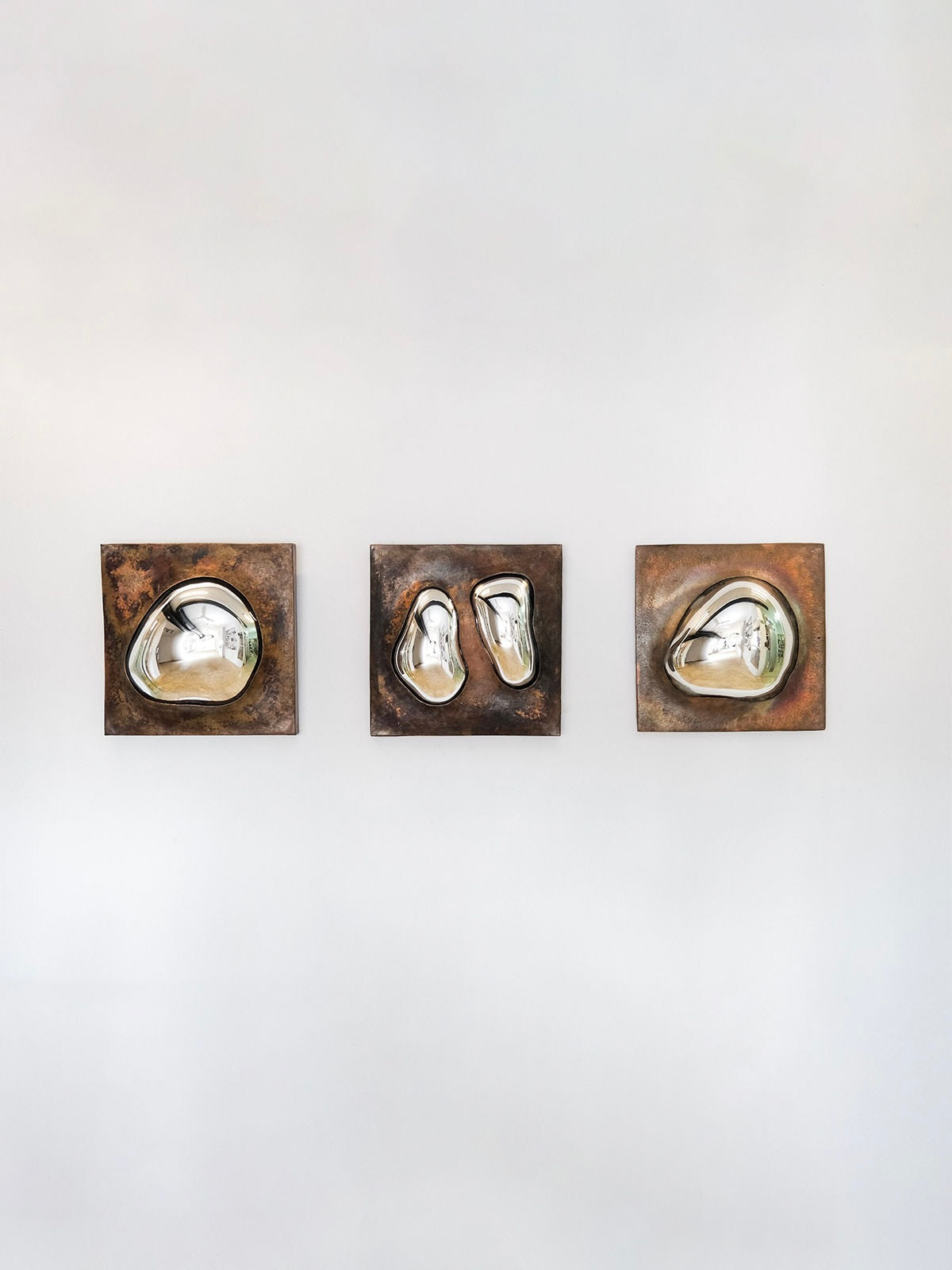 The HOMA "Drop Of Silver On Copper III" features three abstract sculptures mounted on square rust-colored copper frames on a white wall. Each sculpture has a unique, smooth shape resembling liquid droplets, softly reflecting light in a minimal setting.