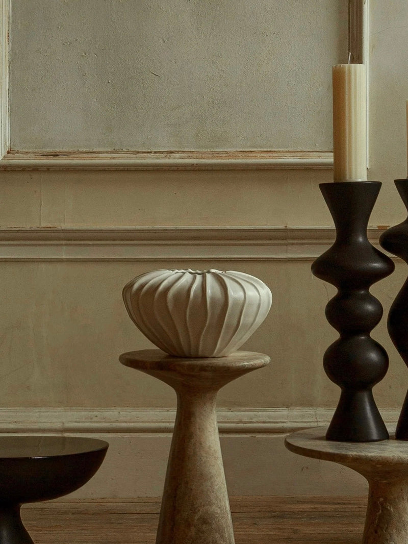 In a minimalist interior, a Wide Cloak Vase by Ransom & Dunn from their Cloak ceramic collection, handmade in Portugal, stands beside two black candlesticks. Meanwhile, a white ribbed planter elegantly rests on a beige pedestal against a textured wall with decorative molding.