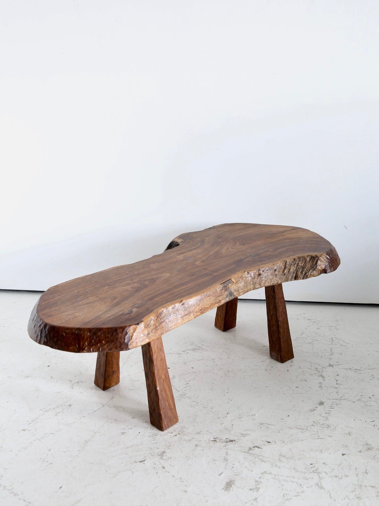 The 'French Folk' Coffee Table by Ancien et Jolie features a rustic, live-edge wooden top with adze-carved details inspired by 1970s French design. Its bold grain pattern and organic edges stand elegantly on three sturdy legs against a light-colored floor and plain background.