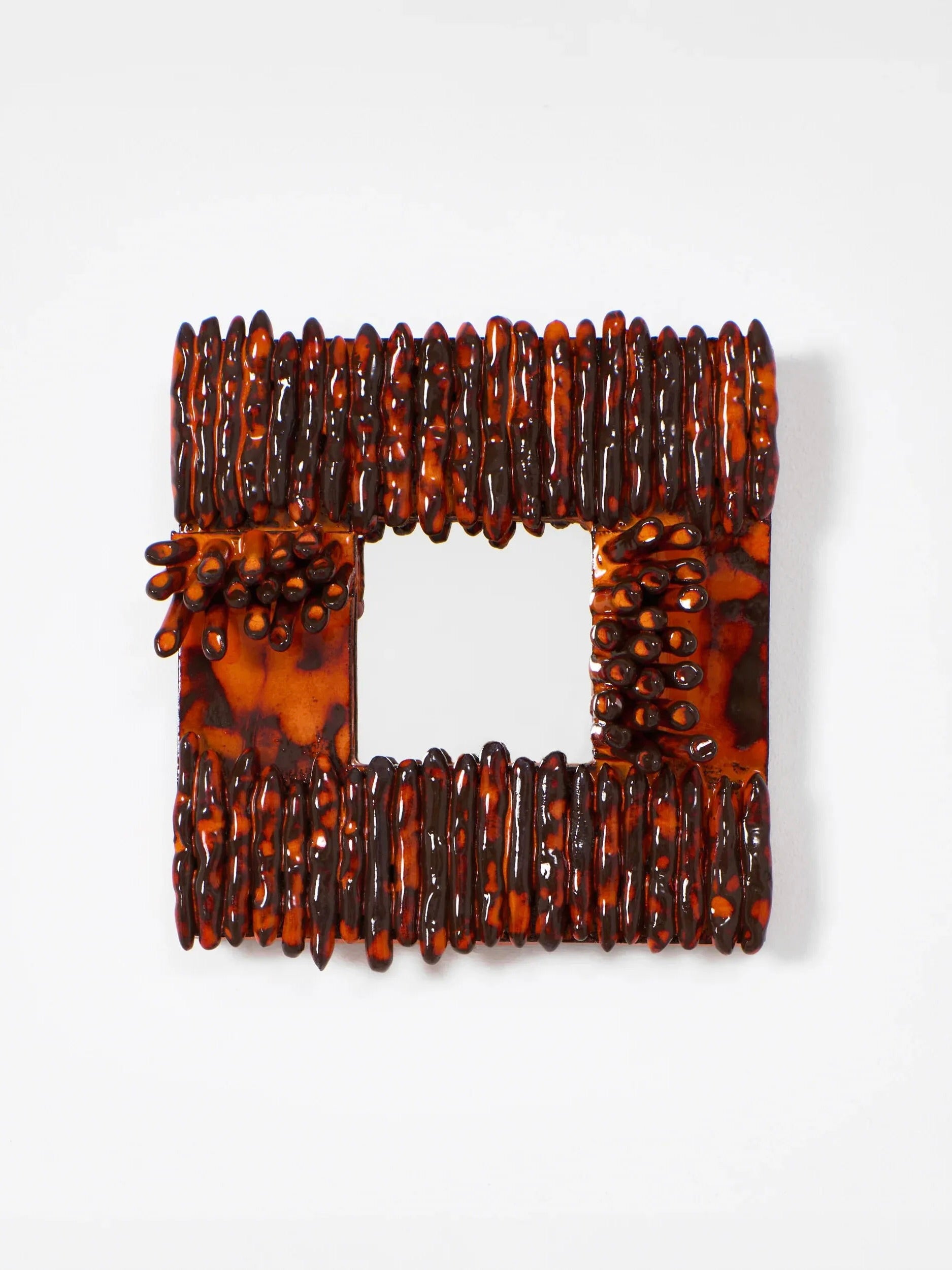 The 'Dedo' Ceramic Mirror by Project 213A is a square art piece with a central hole resembling an artisanal ceramic mirror. It features layered, glossy materials akin to amber with textured surfaces in dark orange and brown, echoing organic, handmade formations for visual dynamism.