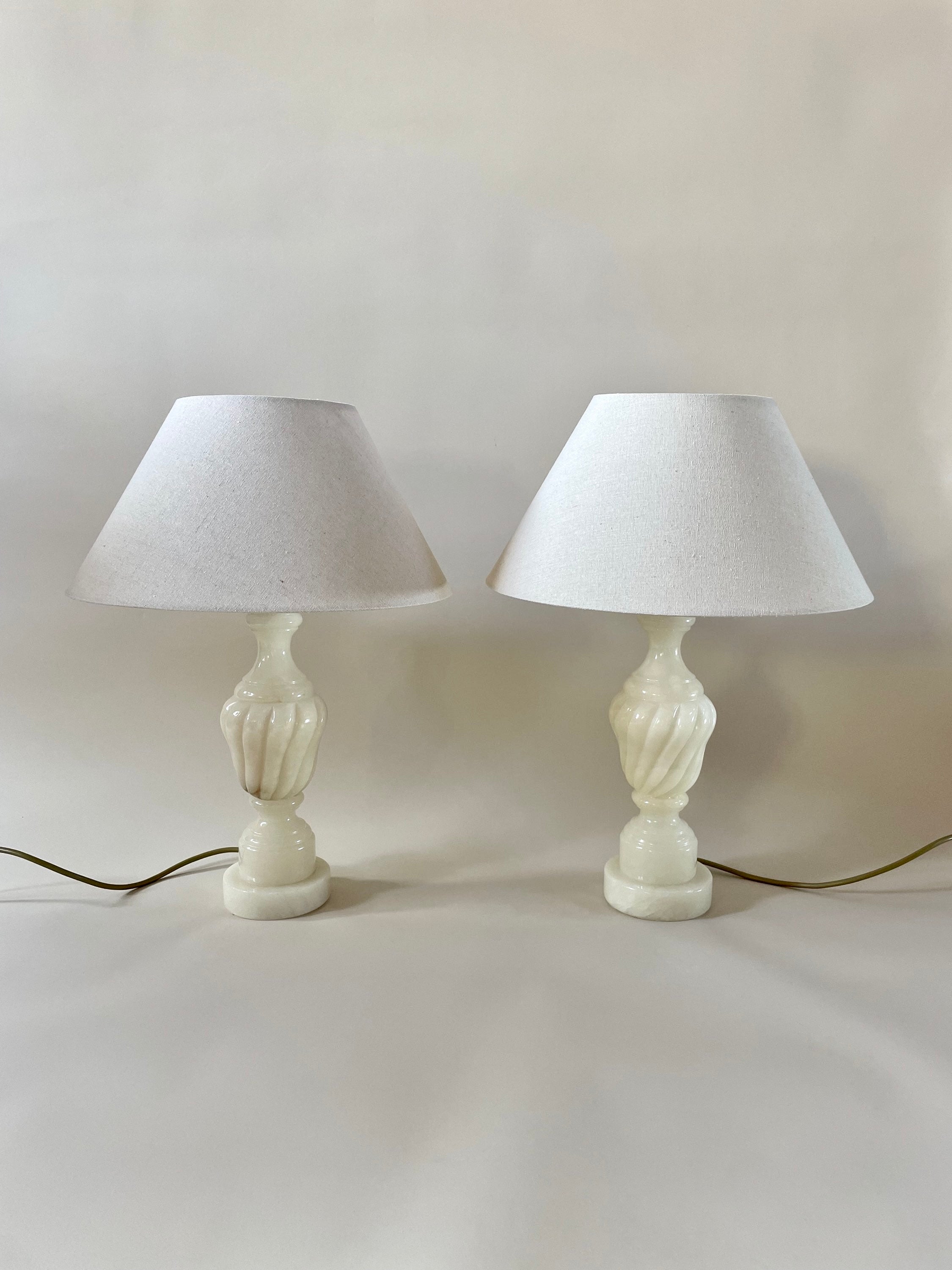 A pair of Maud Vaughan's "Vintage Alabaster Lamps" featuring white shades and bases designed like two clasped hands sit side by side against a plain white background, with antique gold flex power cords visible at the base of each lamp.