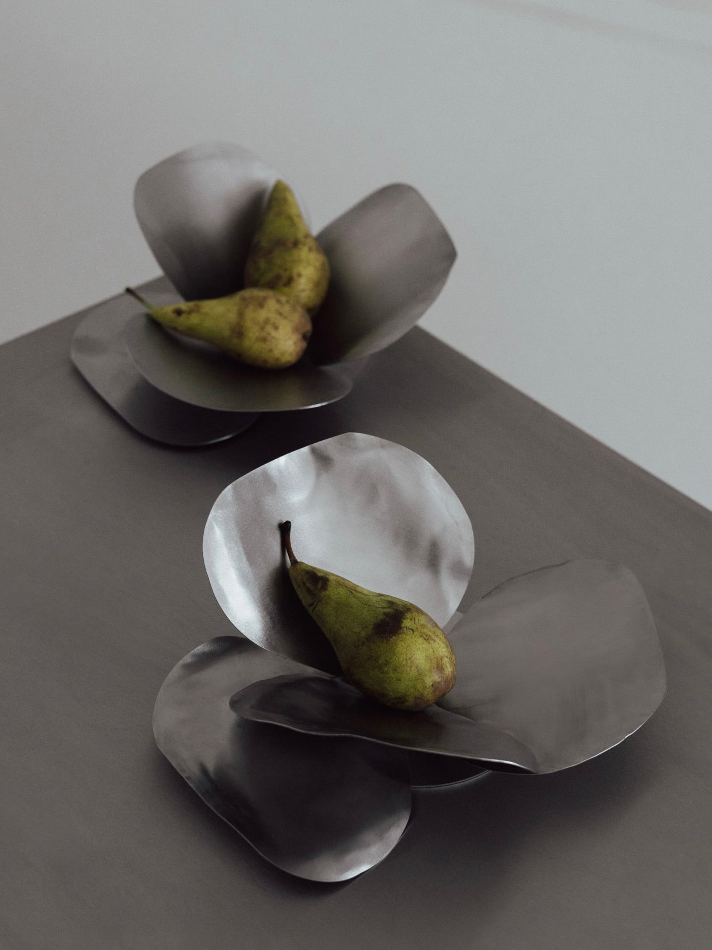 Two Petale Bowl sculptures by The Good Living&Co., featuring metallic, flower-shaped designs, each cradle a ripe pear on a sleek, dark surface. Crafted by artisans from stainless steel, their minimalist design and soft lighting contribute to a modern and artistic atmosphere.