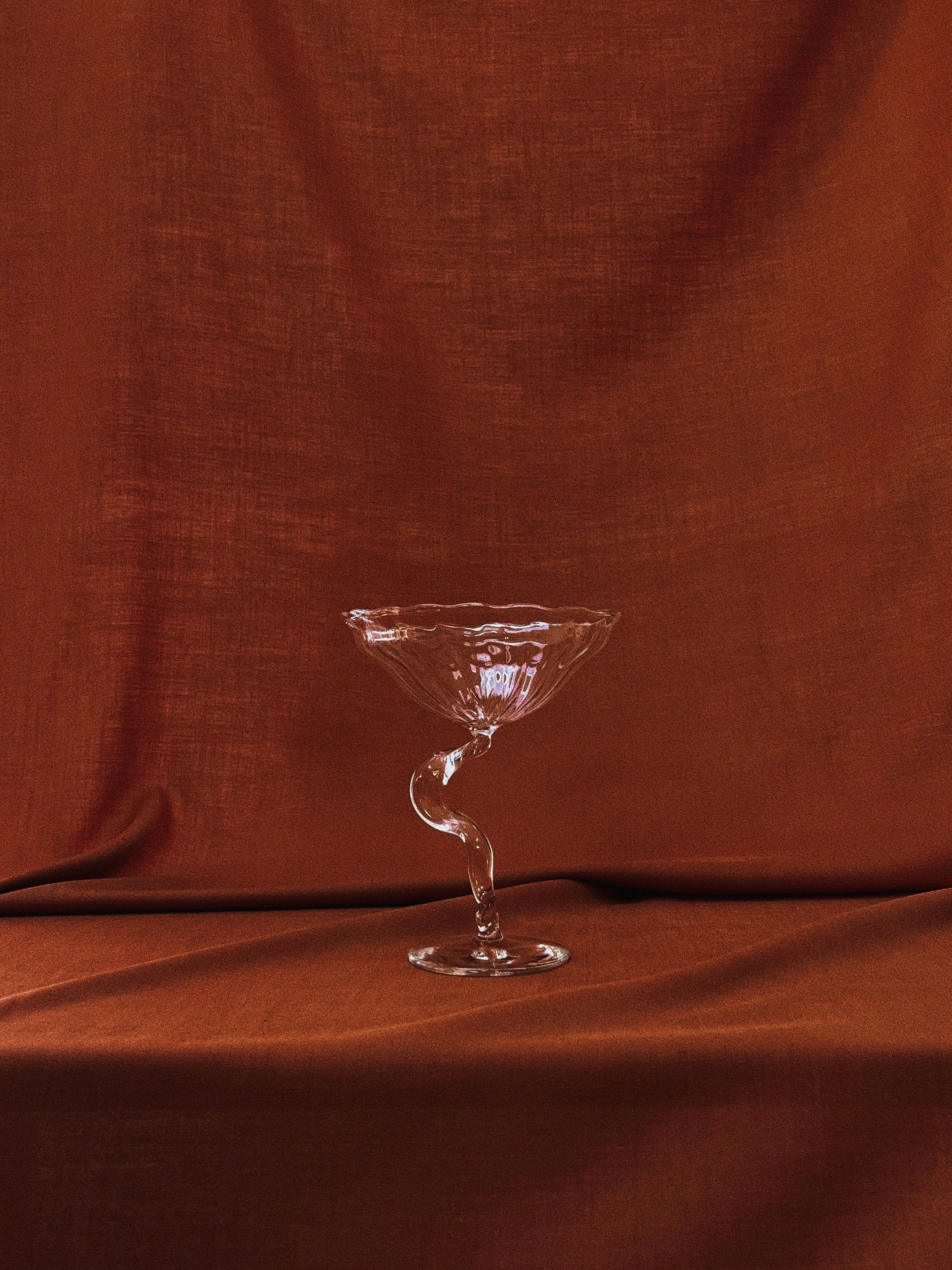 A "Ribbon" Cocktail Coupe from the Hors-d'Oeuvre collection, artfully handcrafted in France with a distinctive twisted stem, is set against a luxurious brown fabric backdrop. This arrangement accentuates the coupe's delicate curves and the fabric's texture, crafting a sophisticated and inviting ambiance.