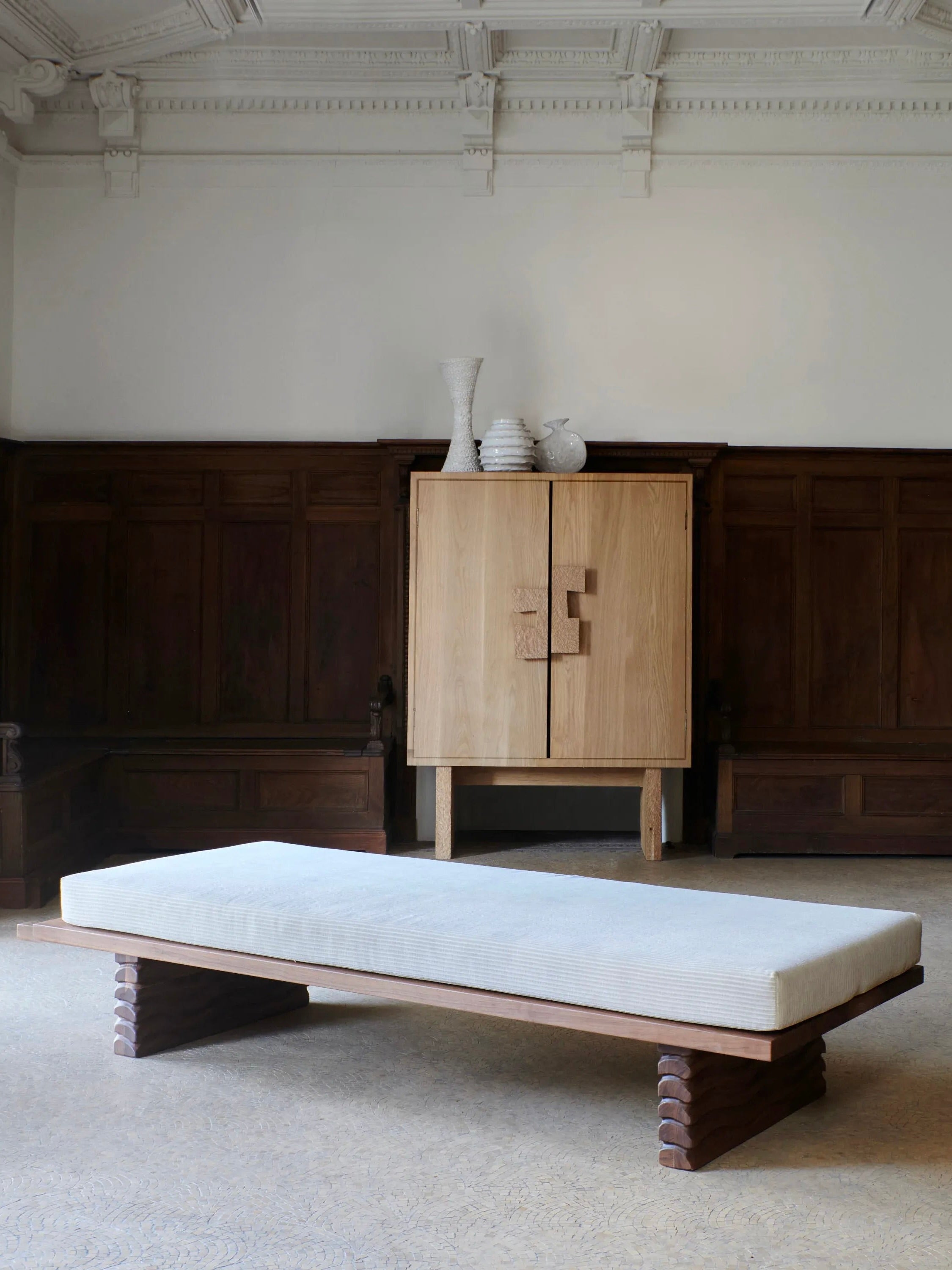 A minimalistic room featuring a Nazaré Daybed by Project 213A with a light-colored cushion, positioned in the center. Behind the daybed, there is a solid walnut cabinet adorned with a collection of handmade ceramic vases from Portugal. The walls have dark wood paneling and ornate ceiling details. The floor is carpeted.