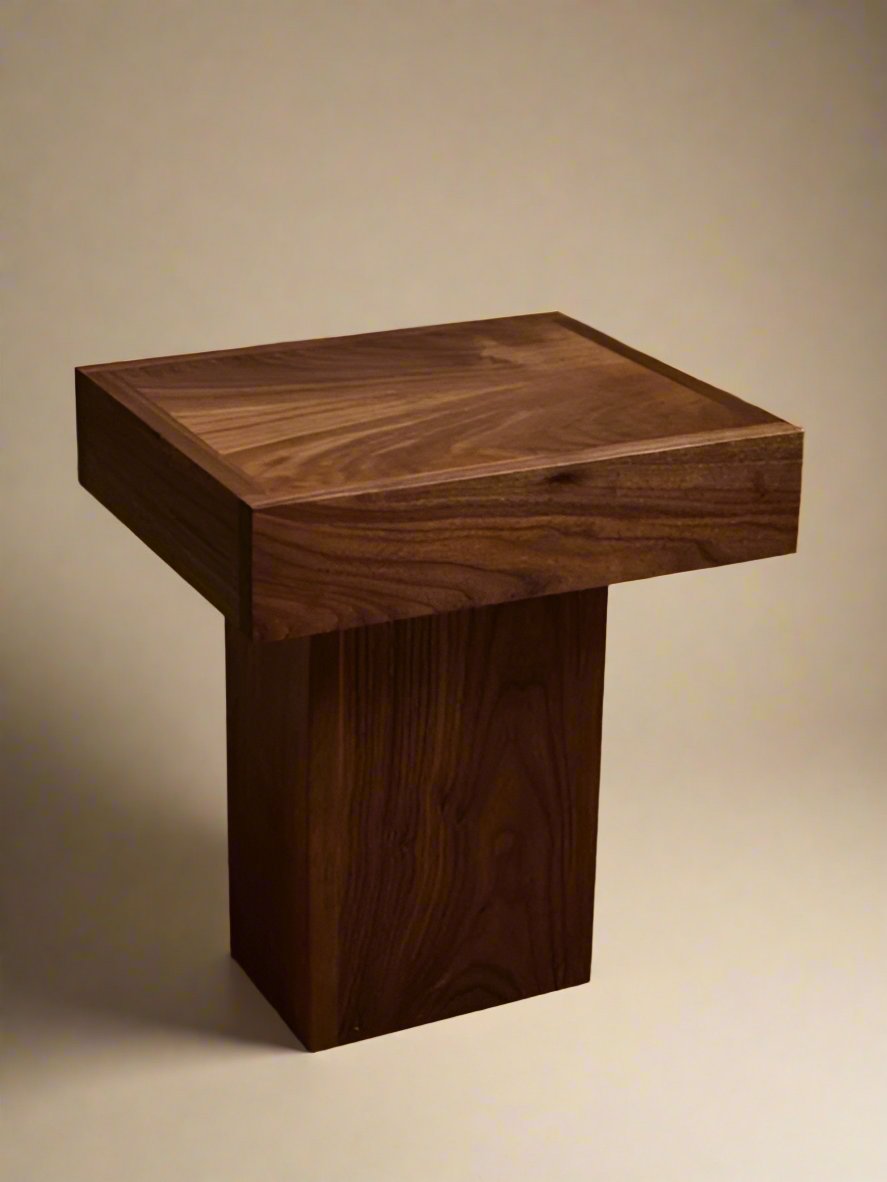 The Jura Pedestal by Ah Um Design Studio is a small, square, wooden side table with a thick top and a single sturdy walnut wood pedestal base. It is placed on a beige carpeted floor with a beige fabric curtain in the background. This handcrafted piece boasts natural grain patterns and a dark brown finish, making it perfect display furniture.