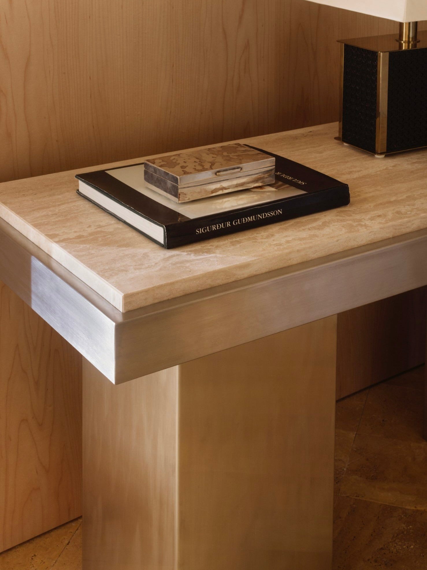 A contemporary Console Table 02 by Studio Vraco, featuring a brushed stainless steel frame and a light-colored surface. The table is adorned with two stacked closed books—one larger and black, the other silver with a textured pattern—alongside a black lamp with a square shade that echoes the elegance of the Bauhaus movement.