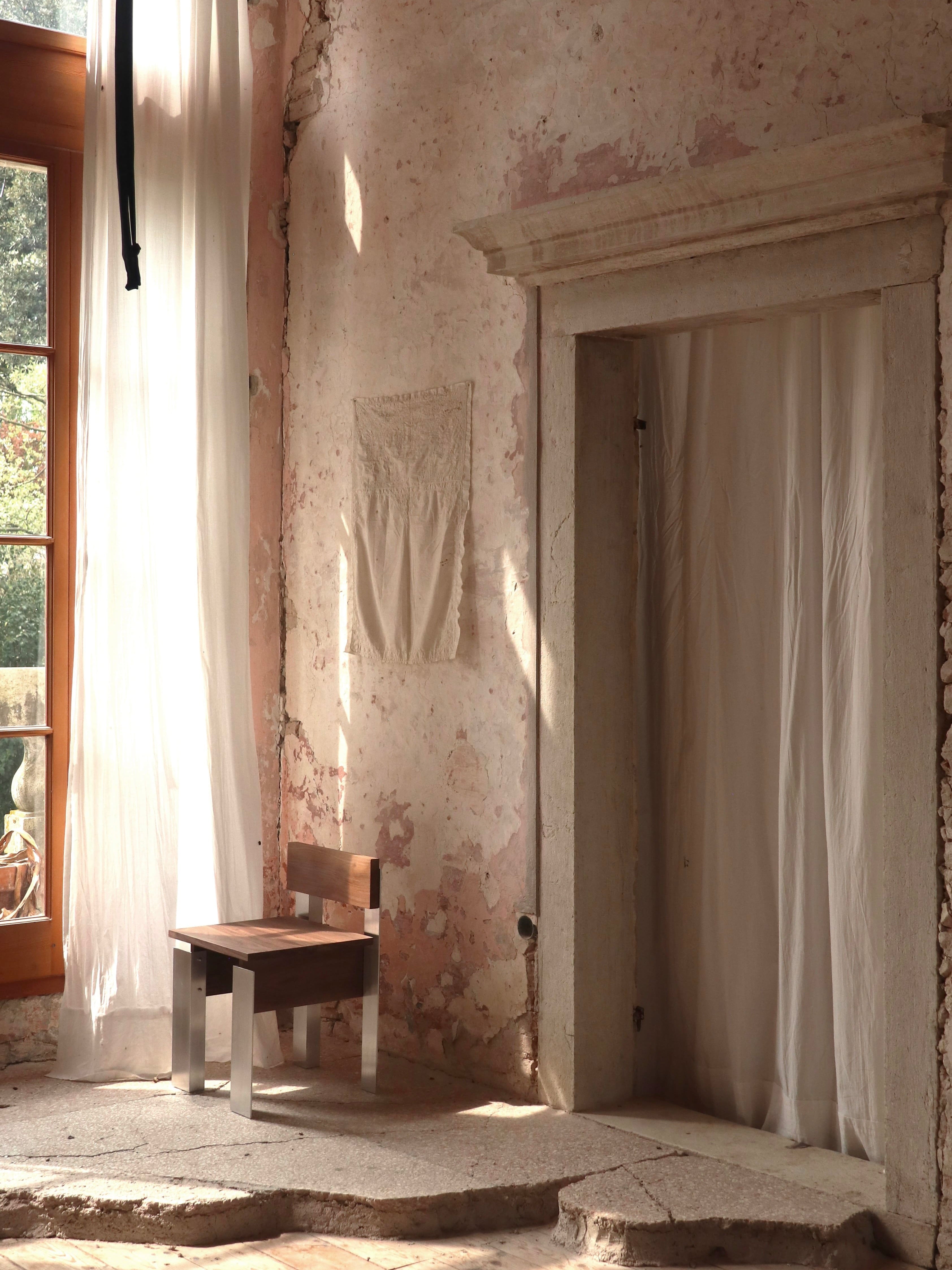 A rustic room features a single walnut Defender Chair by Filippo Andrighetto, nestled by a tall sunlit window. Soft white curtains hang beside the window and doorway, enhancing serenity. The textured, aged walls and stone floor are softly illuminated by gentle light.