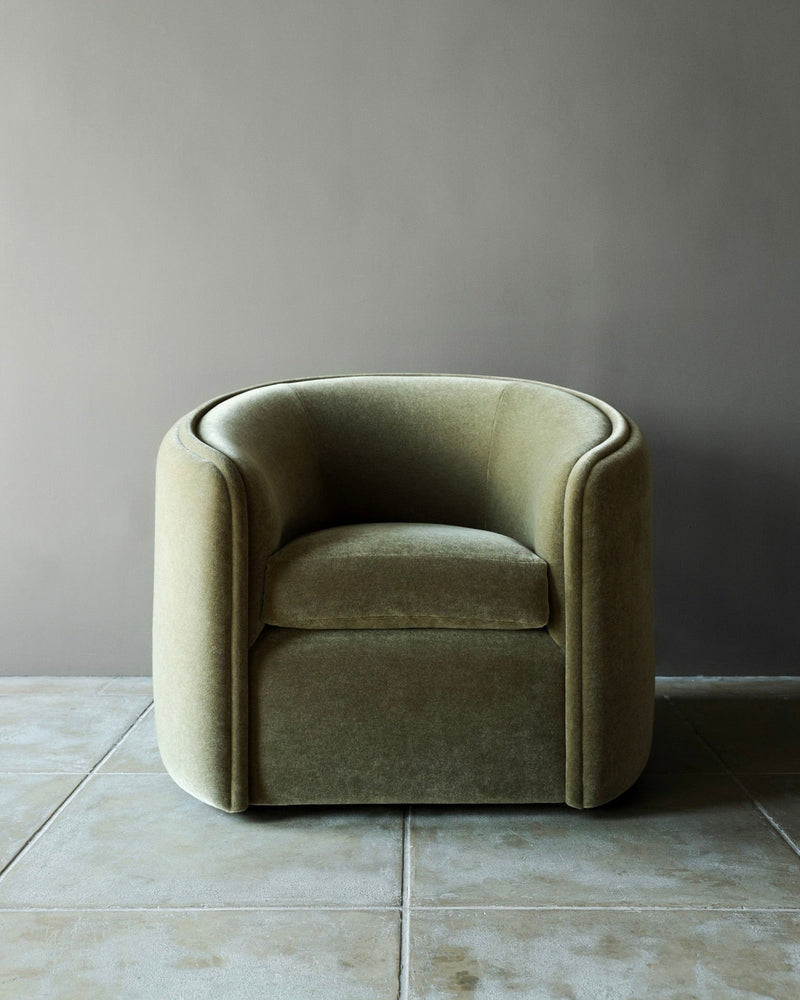The 'DE GAULLE' Mohair Armchair by Studio OSKLO features a modern, olive-green velvet design with rounded edges and a barrel back, highlighting bespoke craftsmanship on large stone tiles against a plain gray wall.