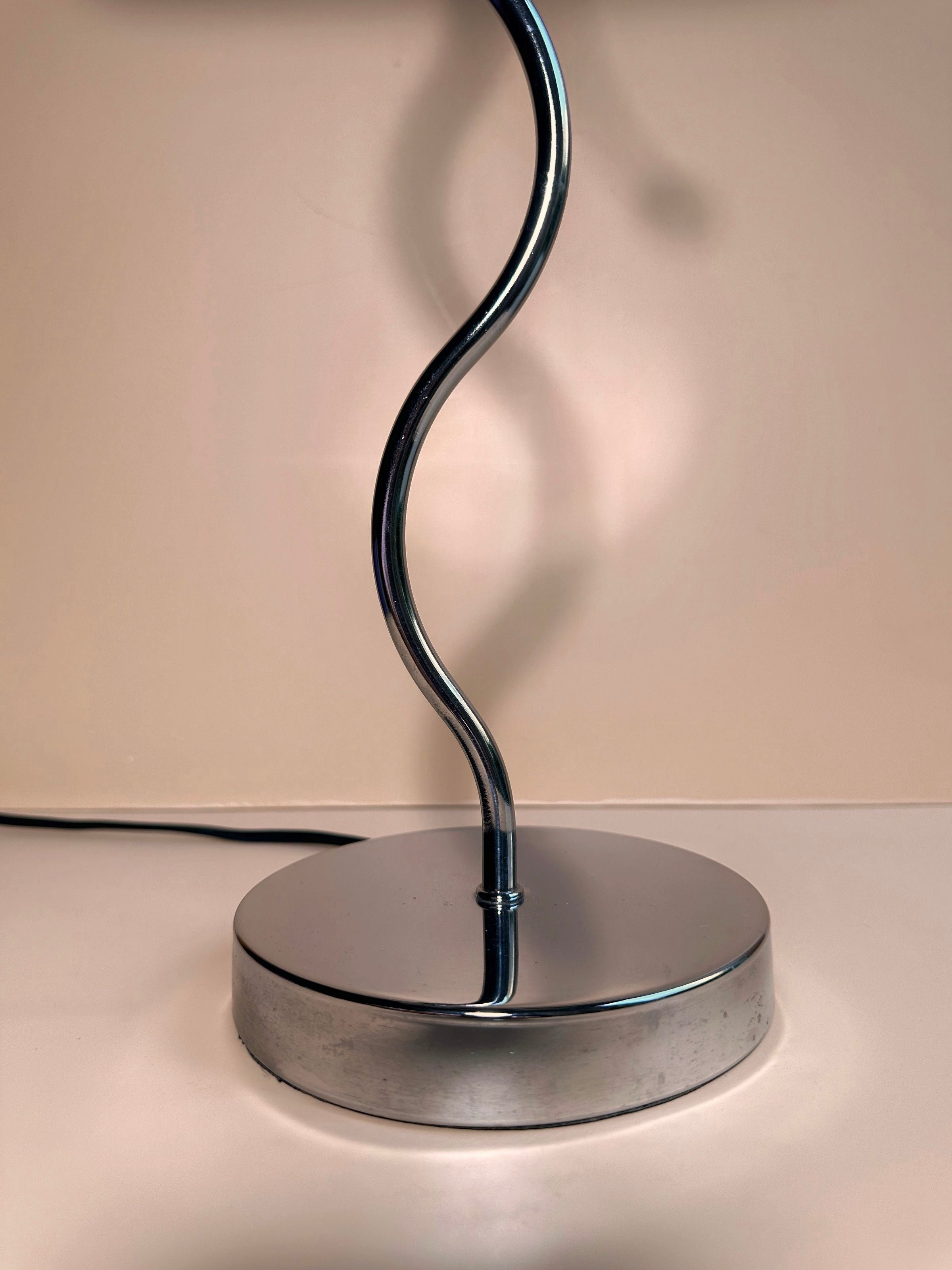 Introducing the Postmodern Wavey Chrome Table Lamp by Dodo Vintage—a contemporary table lamp characterized by a sleek, twisted chrome-plated metal stem mounted on a round, reflective base. The soft beige background accentuates its shiny silver finish and postmodern design, with the lamp's shade not visible in the image.