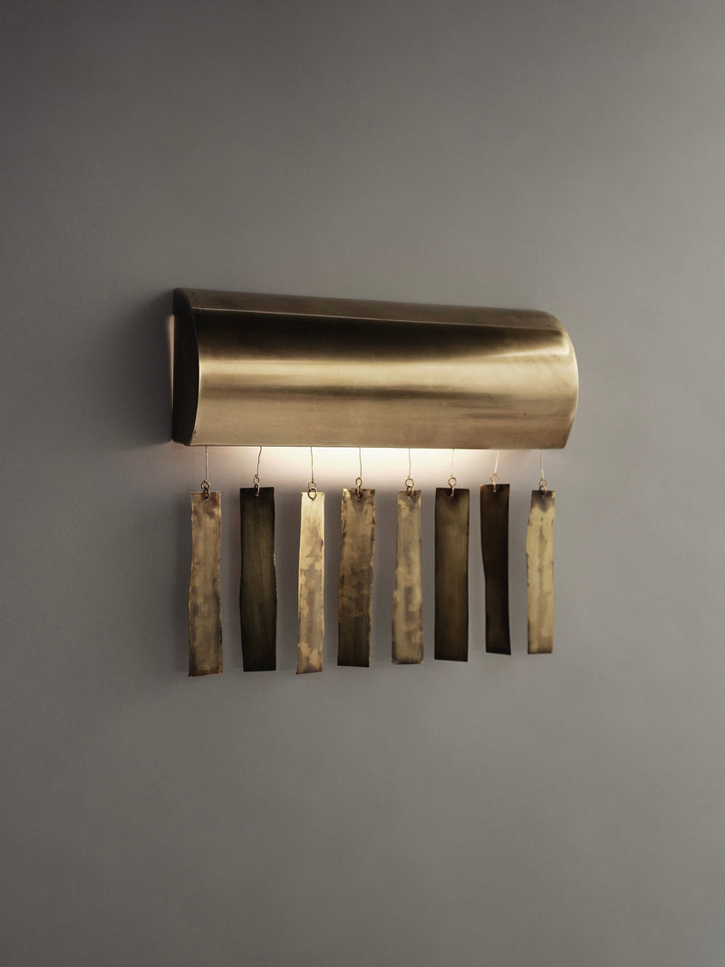 The Ombia Studio Quimbaya Sconce, a handmade modern wall sconce with a polished brass finish, is mounted on a gray wall. Its curved design and vertical metal strips offer contemporary and artistic flair.