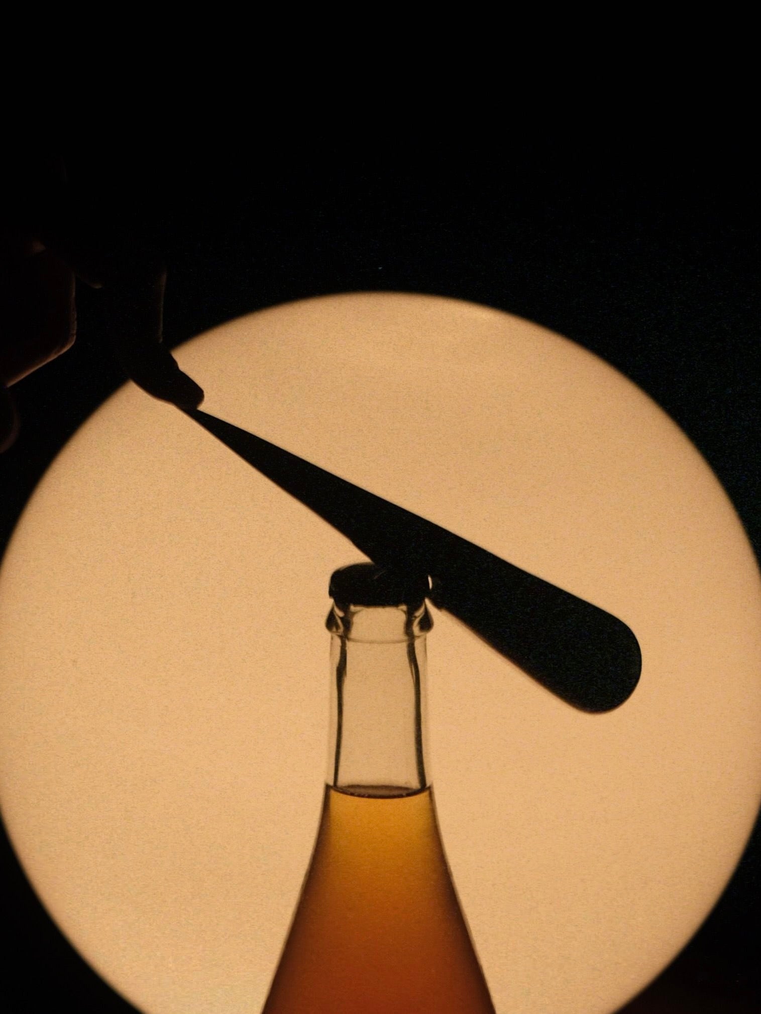 A silhouette of the "Gota" Bottle Opener by Pedro Avila stands with its open top before a circular light source. A hand holds this flat, cast aluminum tool horizontally above the bottle, casting an intriguing shadow against the dark background.